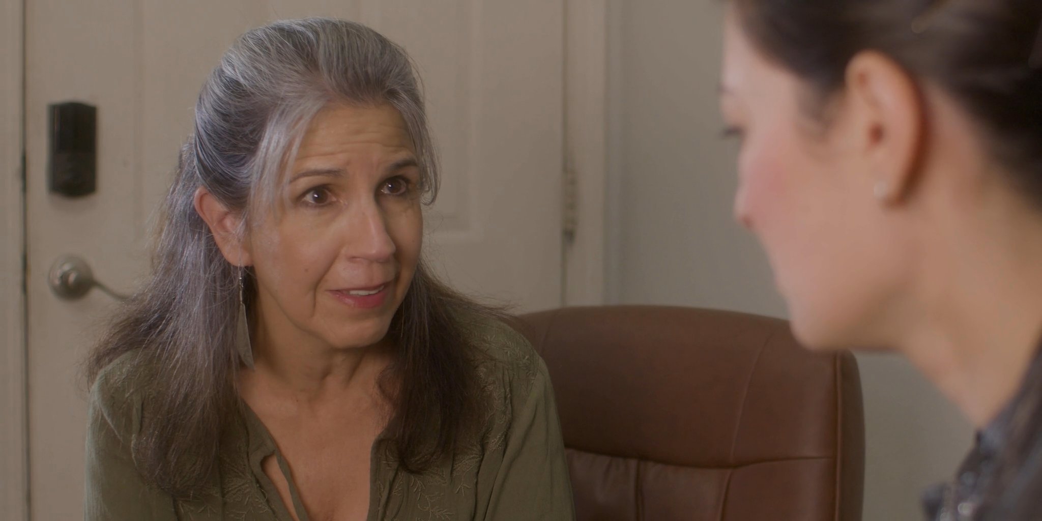 Two women engage in a serious, focused conversation within a professional office setting, exchanging perspectives with visible emotional depth. The dialogue emphasizes attentive listening and supportive interaction between a counselor and a client.