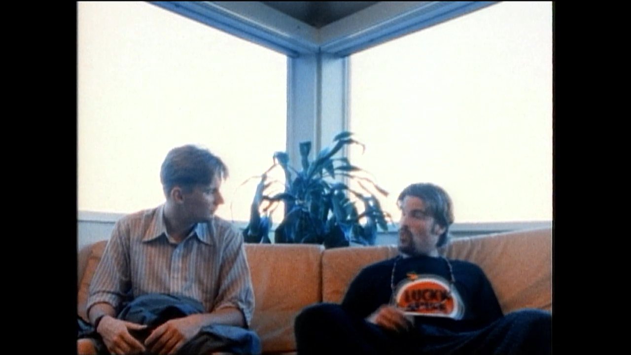 Two men engage in an animated conversation while seated together on a sofa in a brightly lit office setting. The interaction concludes with a cooperative gesture, indicating a consensus or mutual understanding.