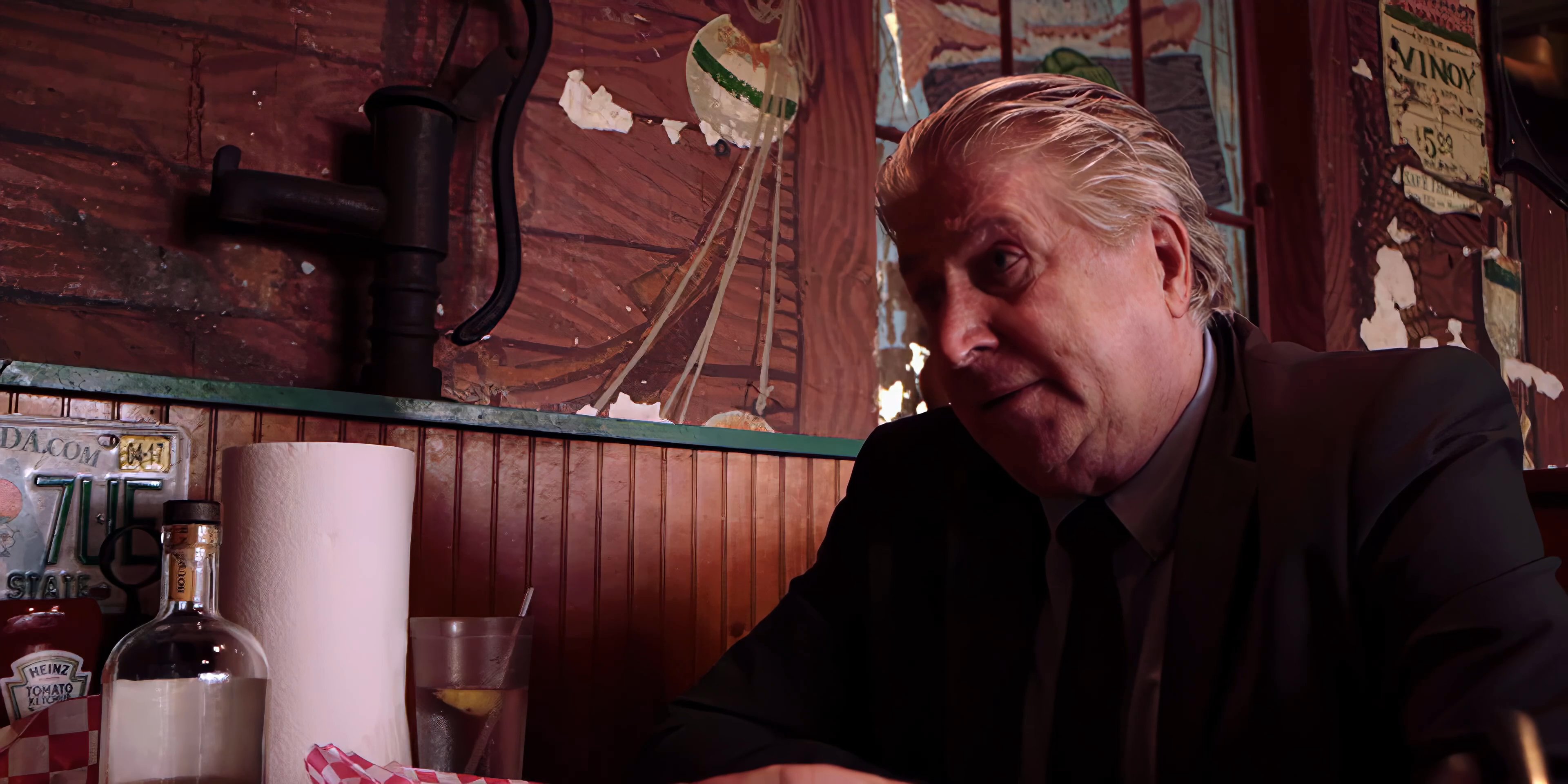 An older man in a suit engages in a serious conversation with a younger man wearing a patterned shirt inside a rustic, vintage-style diner.