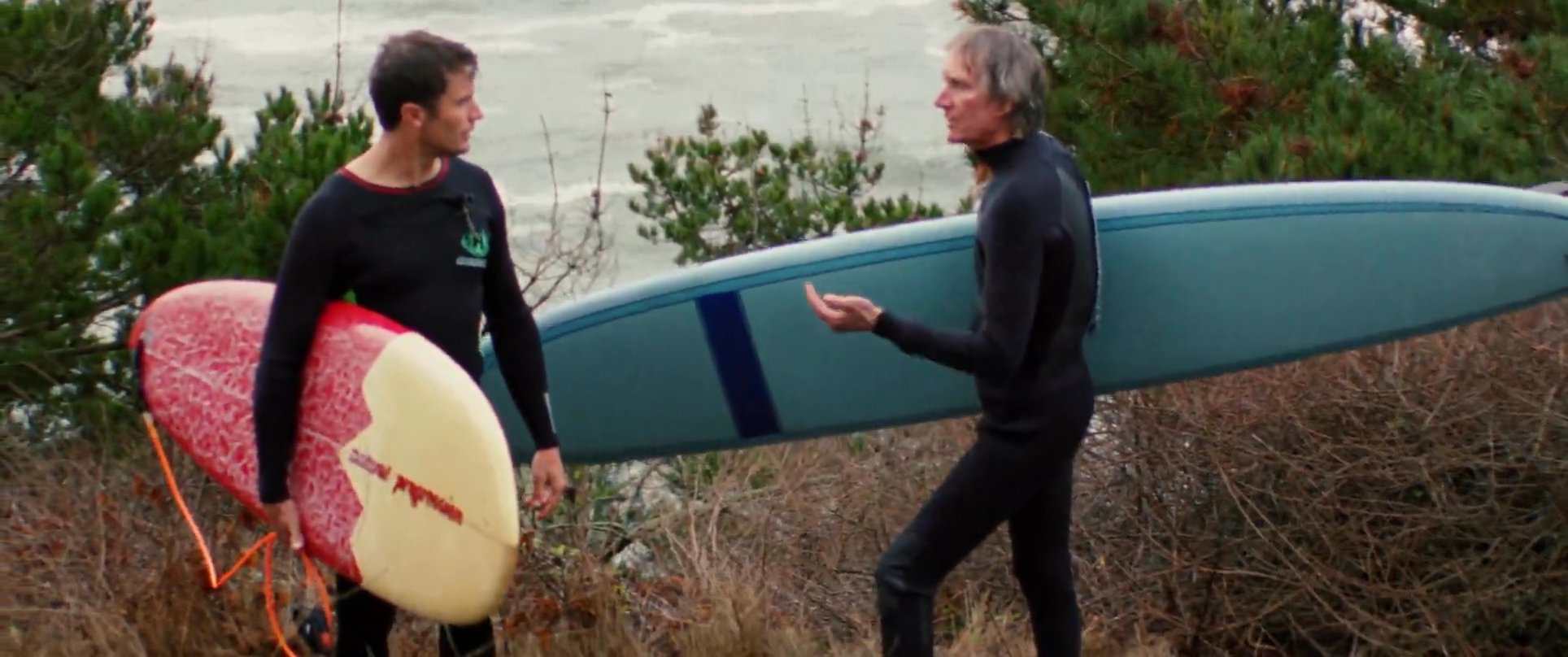 Two surfers dressed in wetsuits discuss conditions while carrying their surfboards across a coastal trail near the ocean. They converse briefly before continuing toward the water.