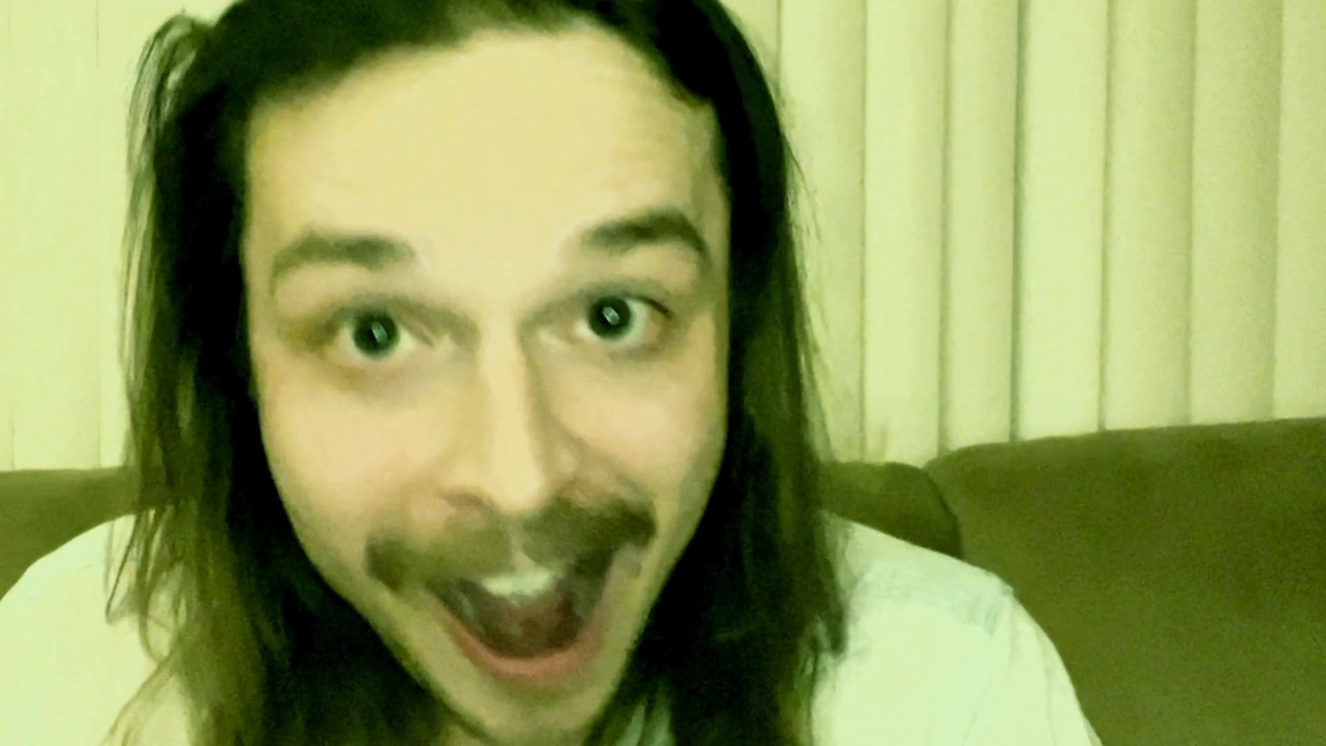 A man with long hair and a mustache engages in an animated, expressive monologue while facing the camera. His facial expressions shift dynamically as he communicates his message.