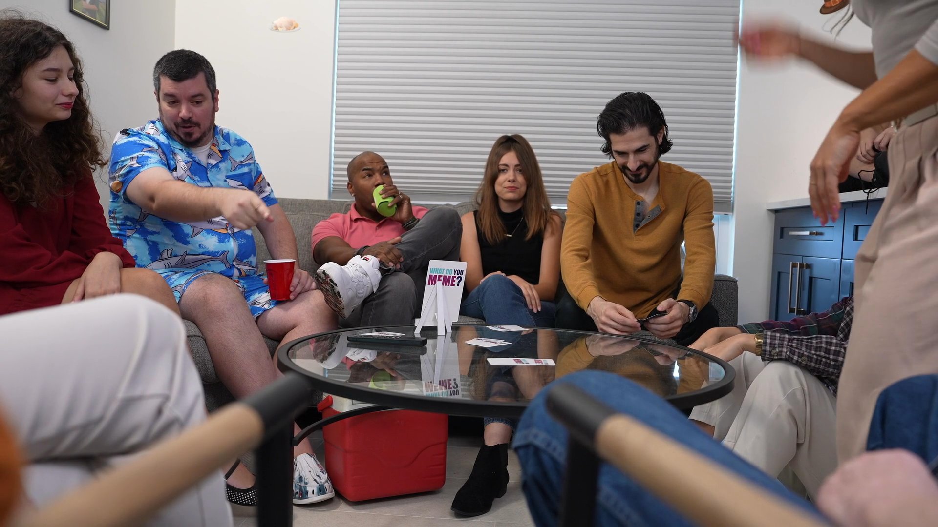 A diverse group of friends participates in a relaxed social gathering in a living room setting. The scene alternates between candid group interactions and individual close-up shots during casual conversation.