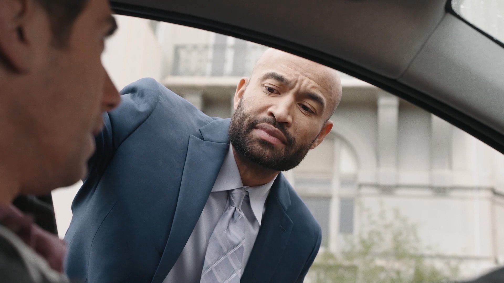 A man in a business suit speaks to a driver seated in a parked car before walking away to leave the lot.