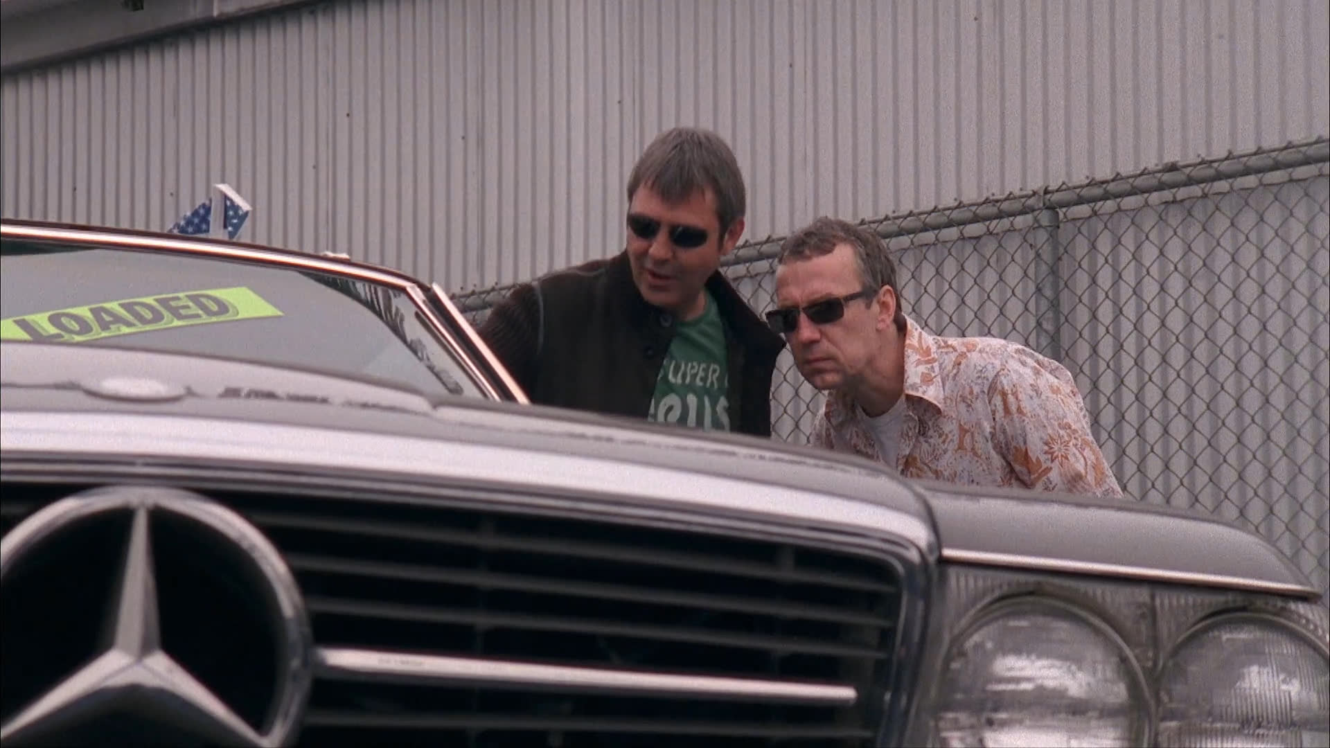 Two men discuss a vehicle purchase at a used car lot before exploring the inventory together. They review various cars on the dealership premises while assessing their options.