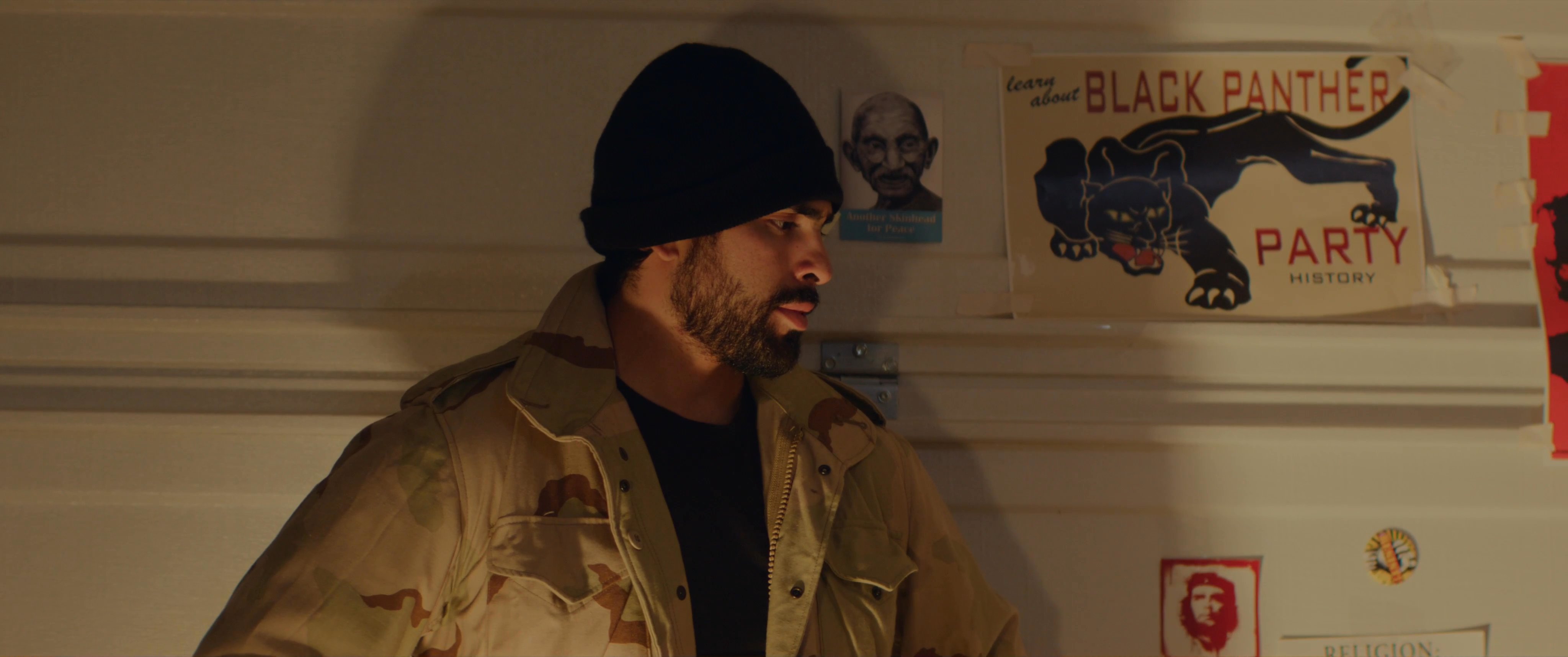 A group gathers in an indoor setting decorated with political posters to conduct an interview or recorded conversation. The participants engage in dialogue while pausing to drink from mugs amidst a film production environment.