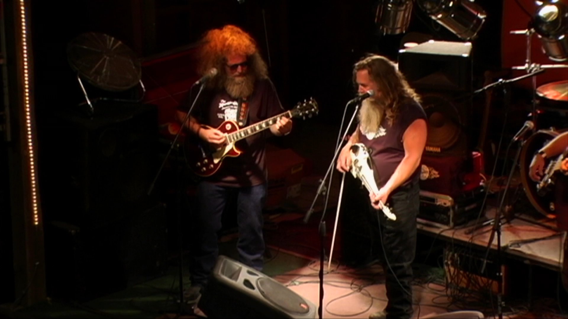 A long-haired, bearded vocalist sings into a microphone while performing on a stage. Beside him, a guitarist accompanies the performance in a dimly lit, professional concert setting.