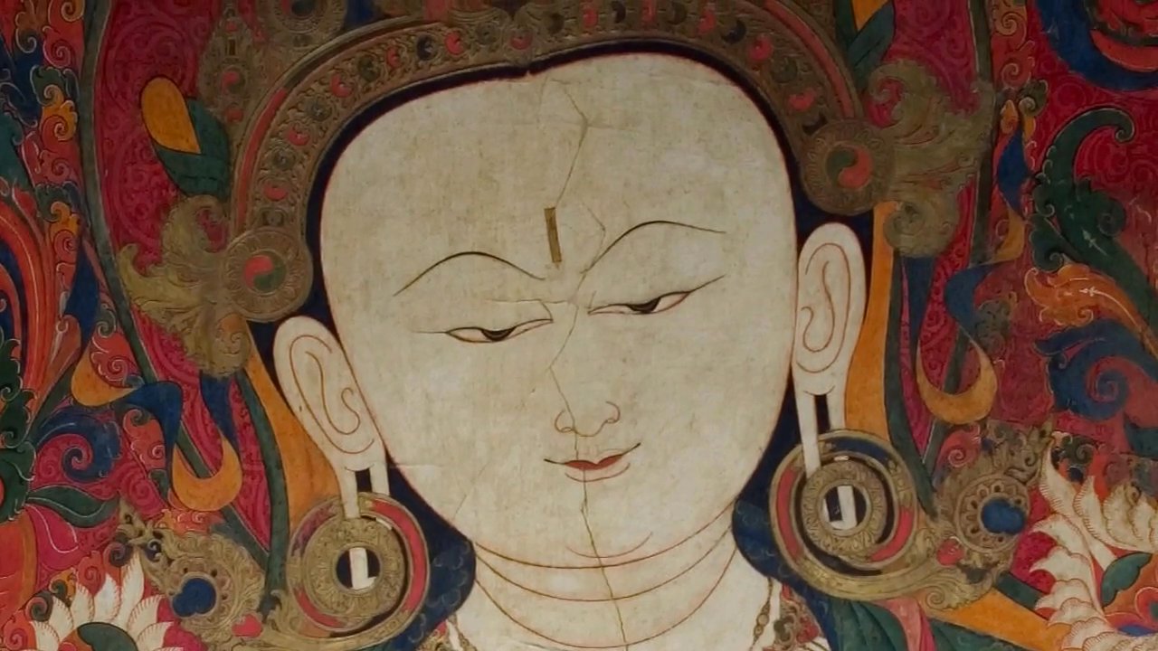 The camera pans across intricate, traditional wall paintings depicting Buddhist deities before settling on an elderly monk standing in prayer within the darkened, atmospheric interior of a Tibetan monastery.