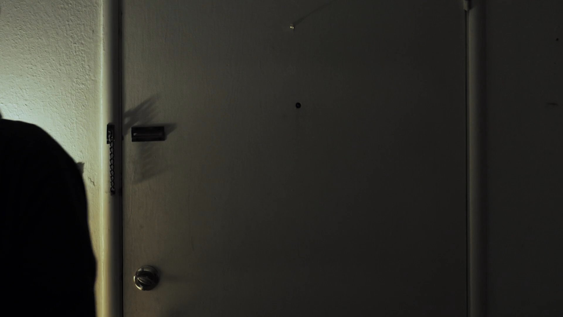 A woman stands by a doorway while a man sits nearby in a recliner, creating a sense of quiet tension. The scene shifts focus to the woman's contemplative and somber facial expressions.