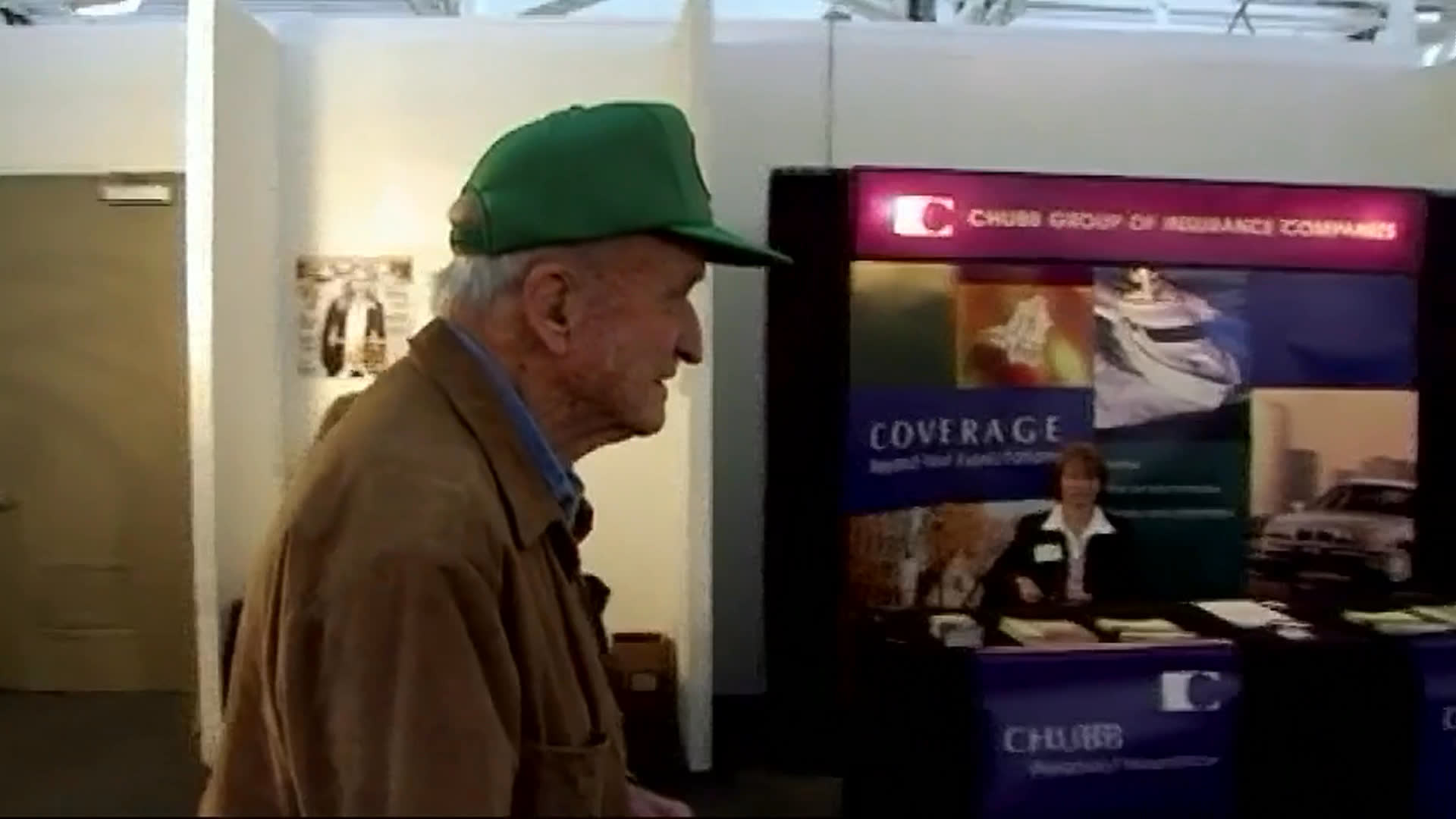 An older man in a green cap points out a large branch-like sculpture and discusses its roughly $20,000 price. He continues through the gallery, viewing artwork and speaking with other visitors.