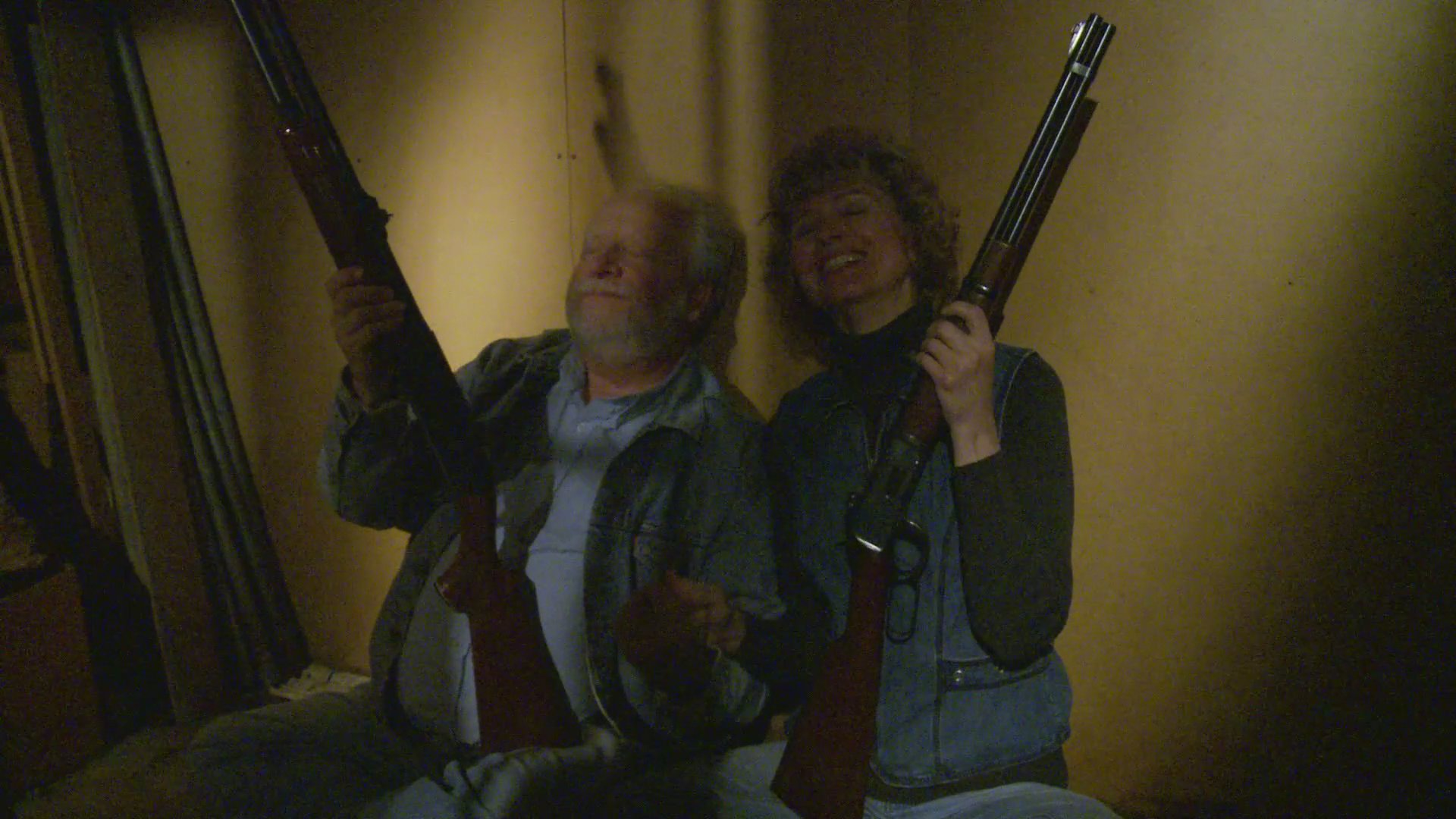 An elderly couple sits together inside a dimly lit room, holding rifles while maintaining a vigilant watch for potential intruders. The scene transitions from a visual double exposure with a police officer at a doorway to the focused pair waiting defensively in their home.