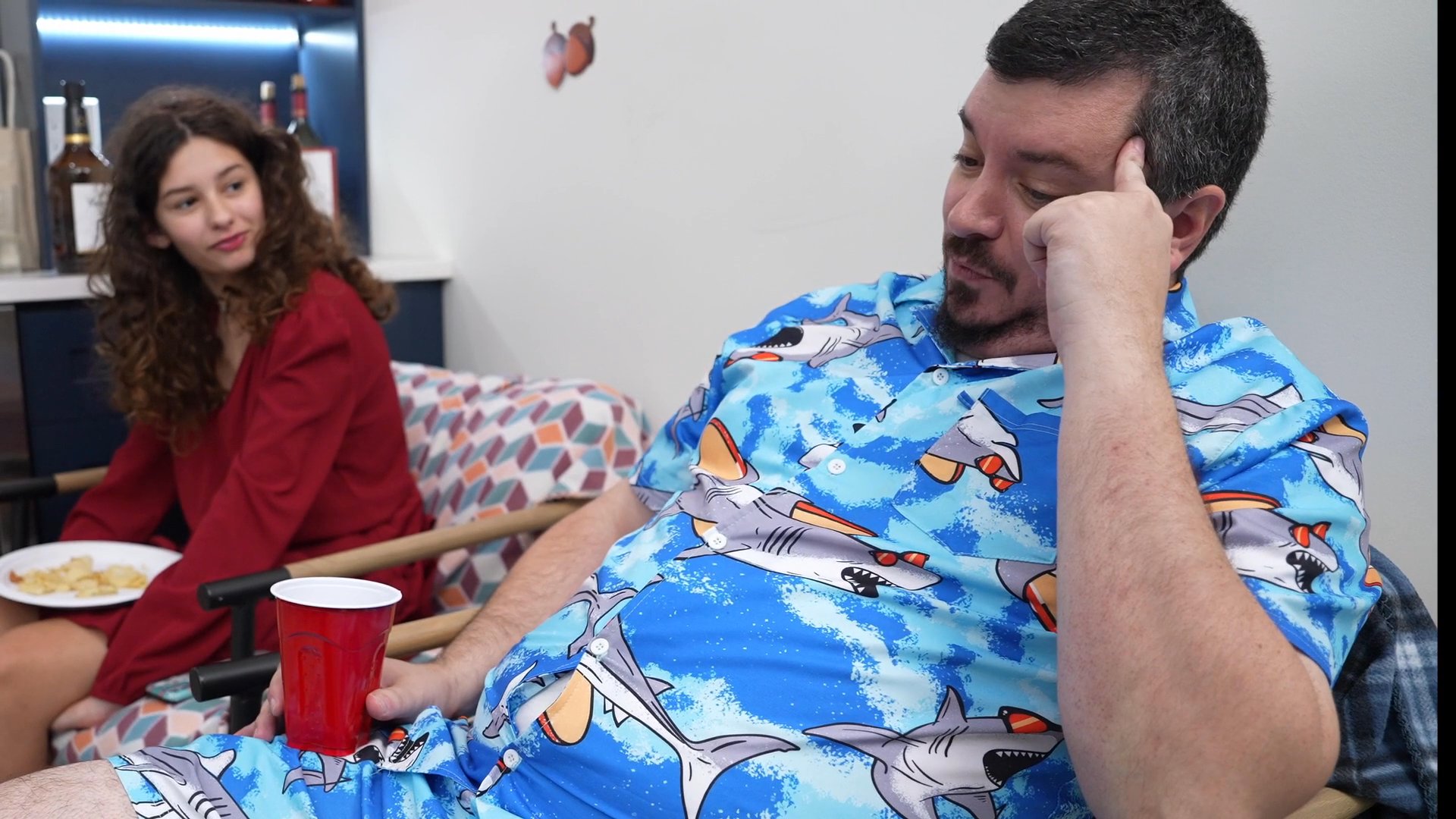 A man wearing a patterned blue shirt engages in a relaxed conversation with a woman in a red outfit while sitting in a living room. The scene transitions to domestic activity as other individuals move through the hallway space.