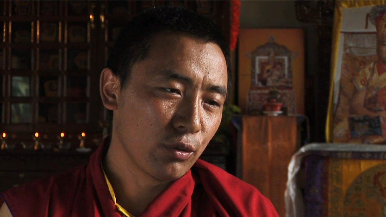 Monks in traditional ceremonial attire perform ritual dances and music at a Himalayan monastery while a speaker recounts the significance of the cultural gathering.