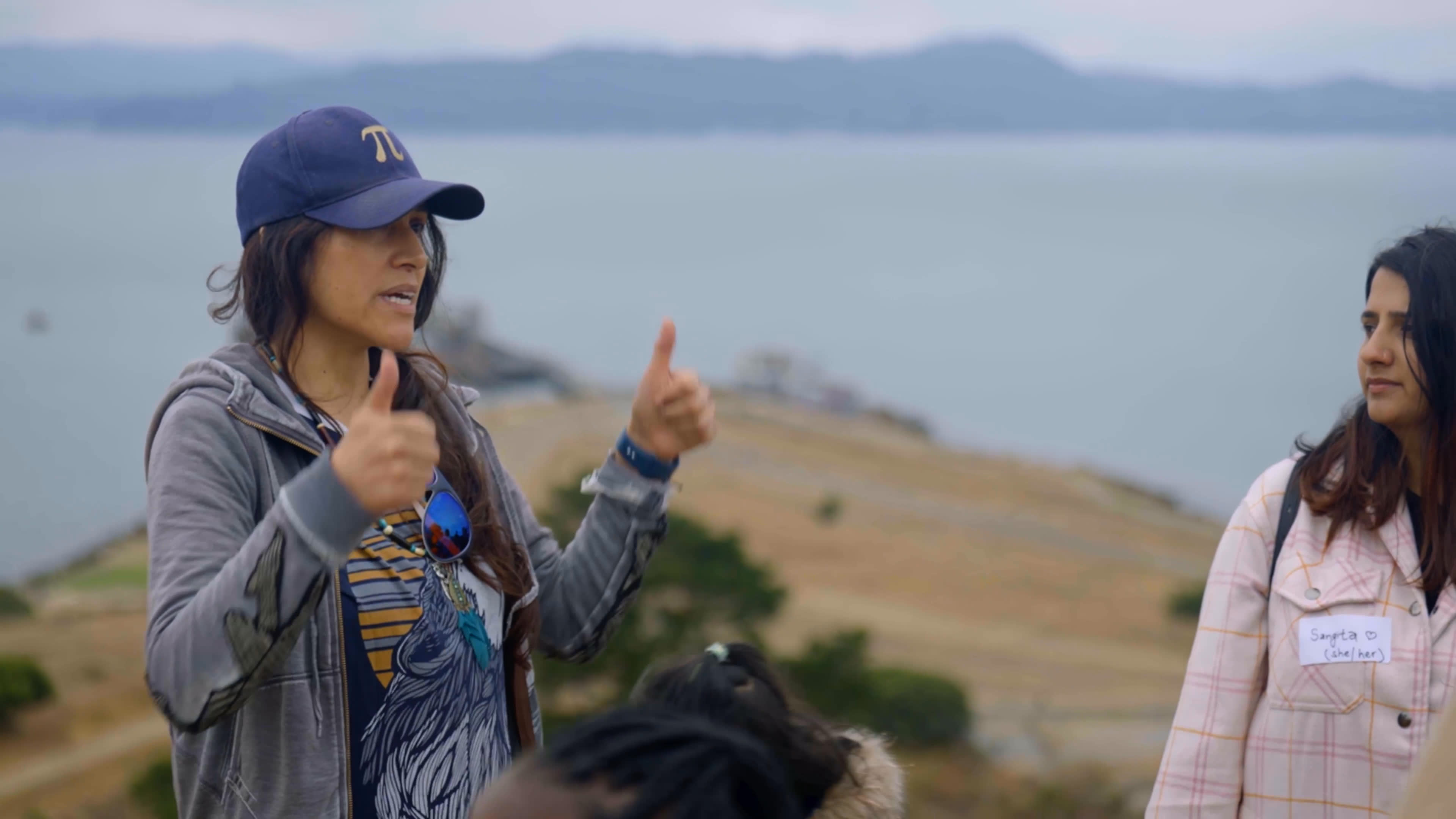 A woman explains how air pollution from a nearby industrial facility affects residents, citing lung cancer, asthma, and heart disease. She gestures while speaking beside a shoreline as another woman and community members listen.