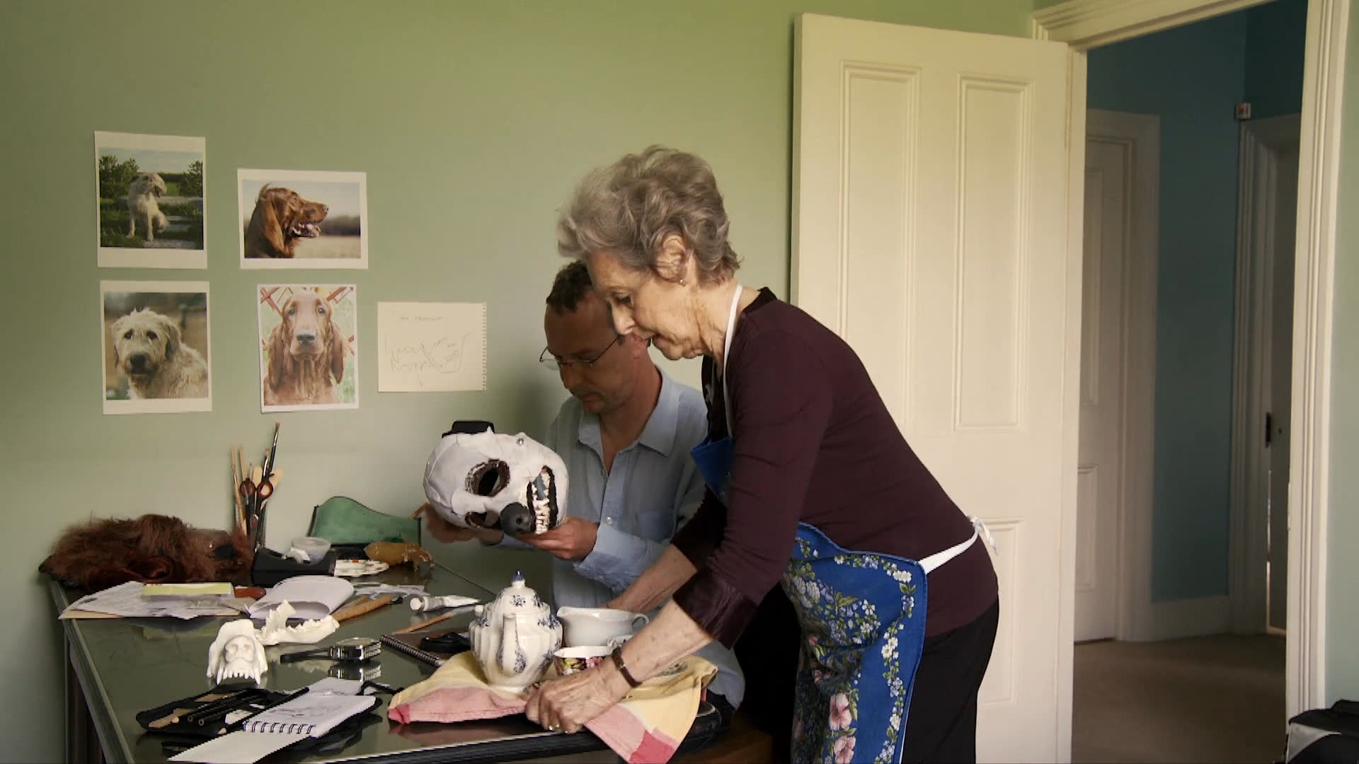 A man sets up camera equipment for a project involving a skull-like mask while a woman prepares tea at a desk. The workspace features various reference images and production props, illustrating a collaborative creative environment.