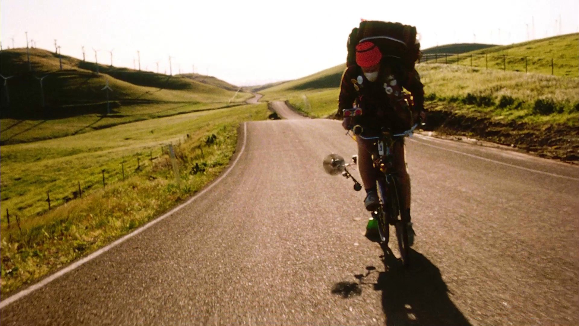 A cyclist wearing an orange beanie and protective mask rides along a rural road, hauling gear decorated with activist-themed buttons. The traveler continues uphill against a backdrop of rolling green hills.