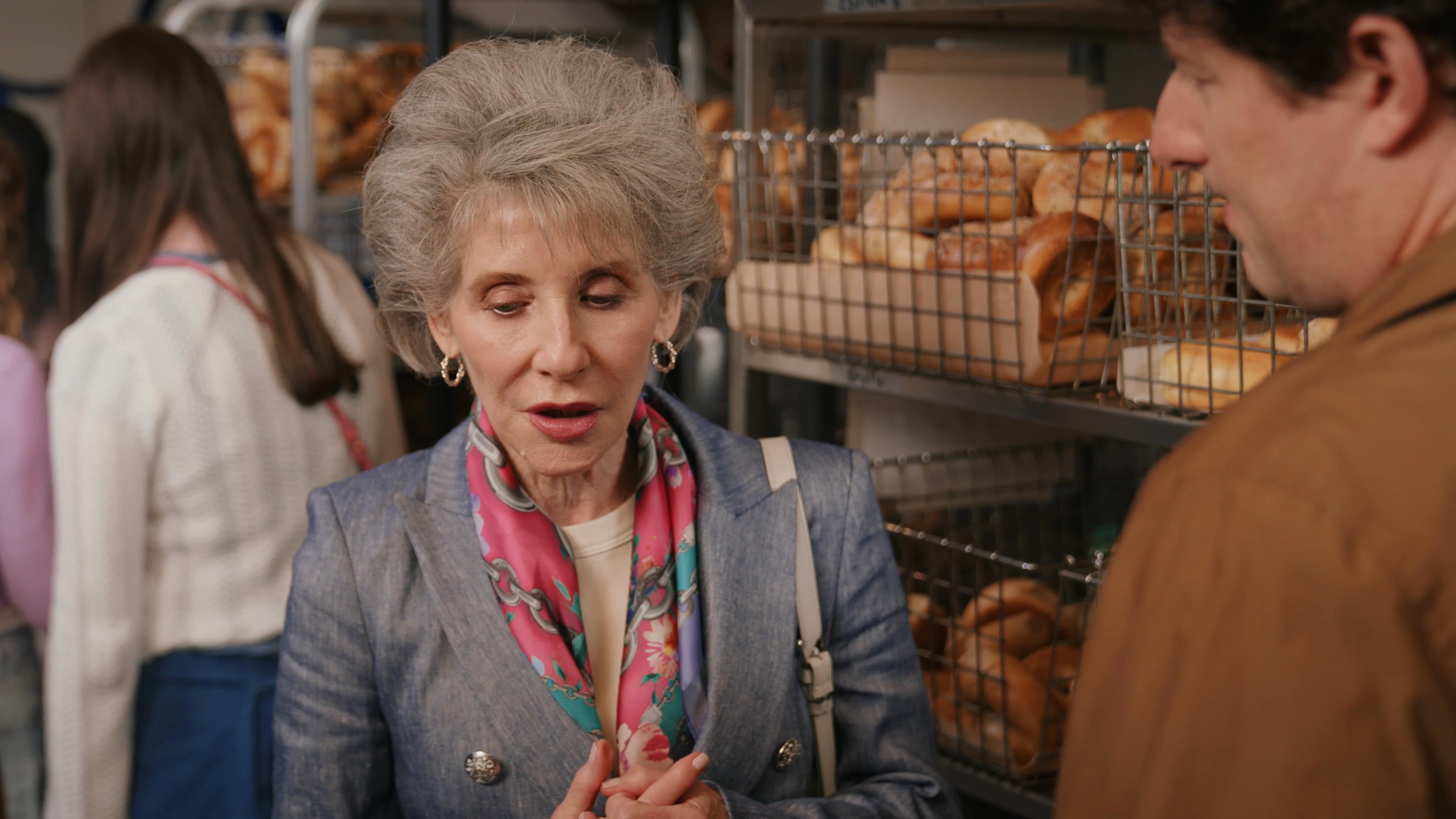 An older woman engages in a serious conversation with a male bakery employee amidst shelves of freshly baked goods. The two exchange viewpoints in a retail setting while discussing specific details.