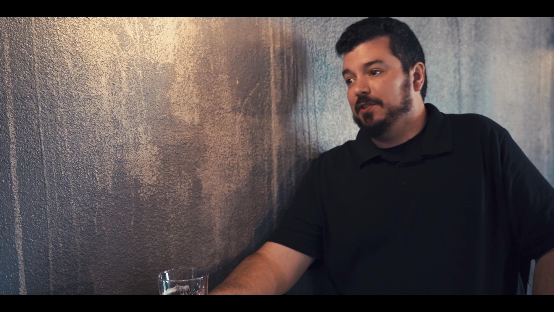 Two men sit at a table in a dimly lit bar, engaged in an earnest, animated conversation.