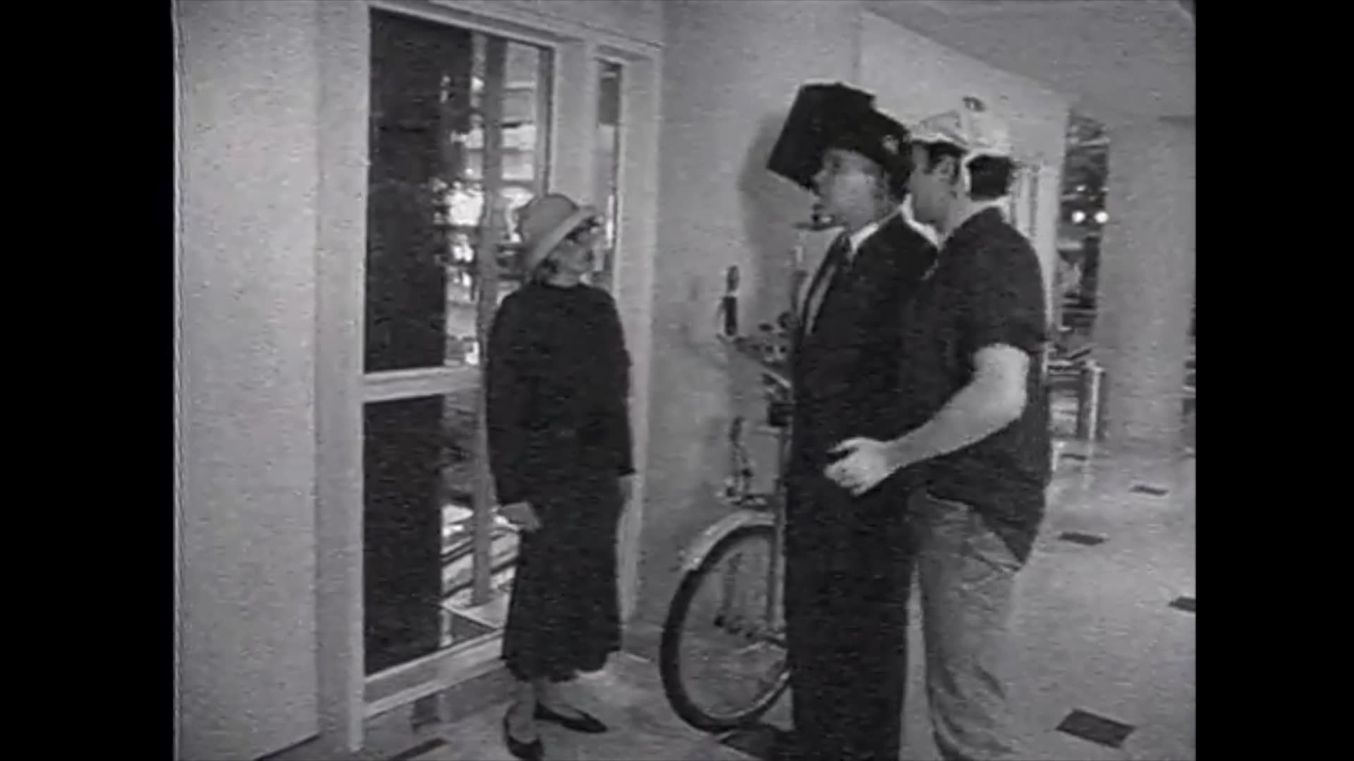 A woman in a cloche hat engages in a stylized conversation with two men, one of whom is dressed in a formal suit and top hat while standing next to a bicycle in an austere, monochrome setting.