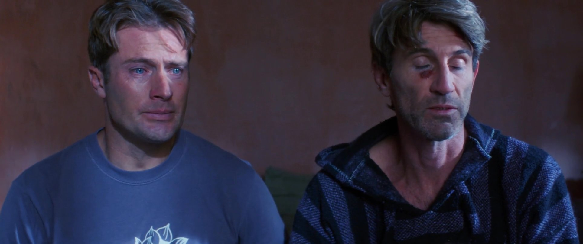 Two men engage in an intense and tearful conversation in a dimly lit bedroom as they process shared grief or conflict. The exchange highlights a moment of vulnerability and emotional support between the pair.