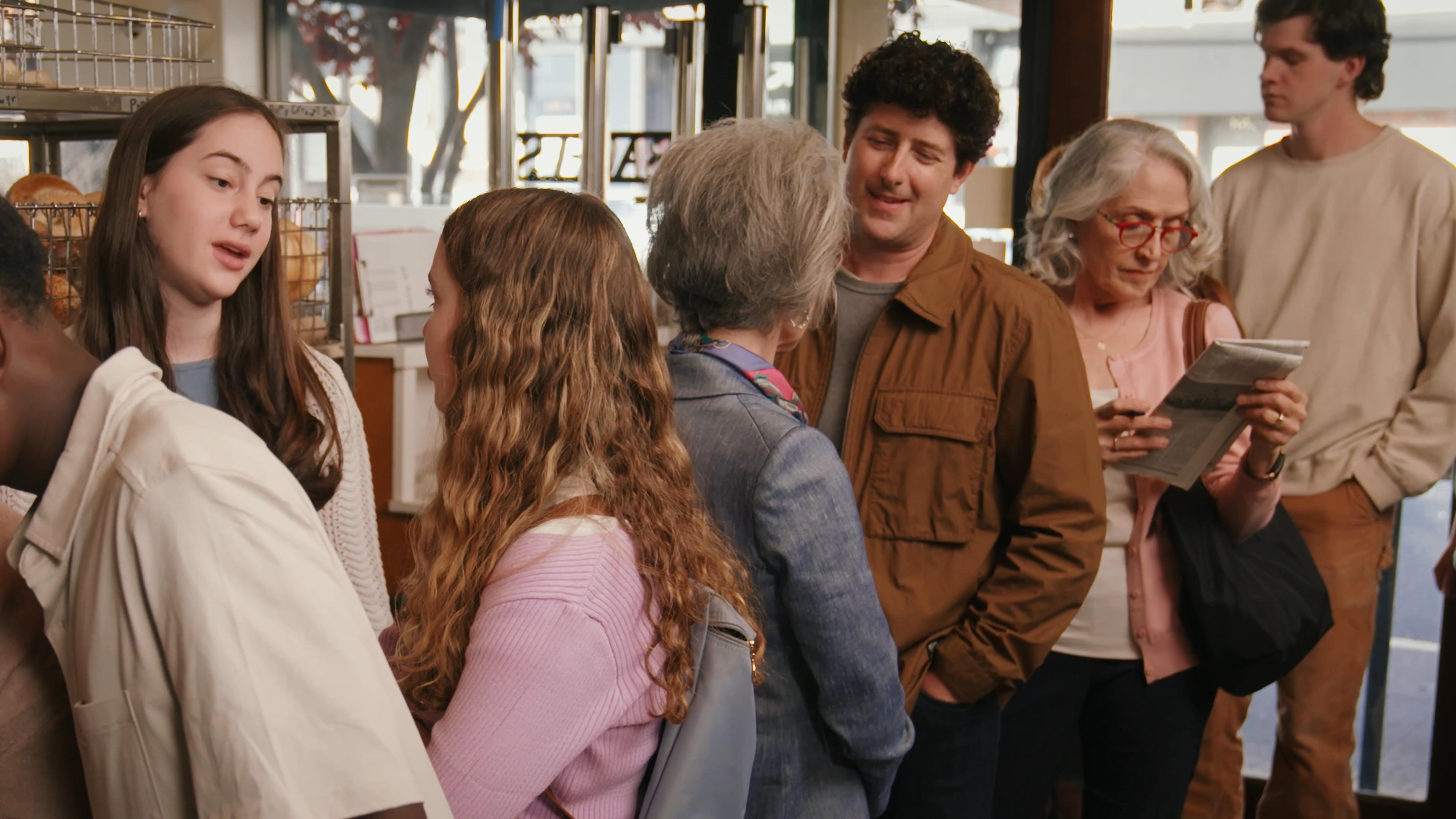 An older woman engages in a friendly conversation with a younger man while standing in a busy bakery shop. The interaction concludes with a warm, affectionate gesture between the two individuals.