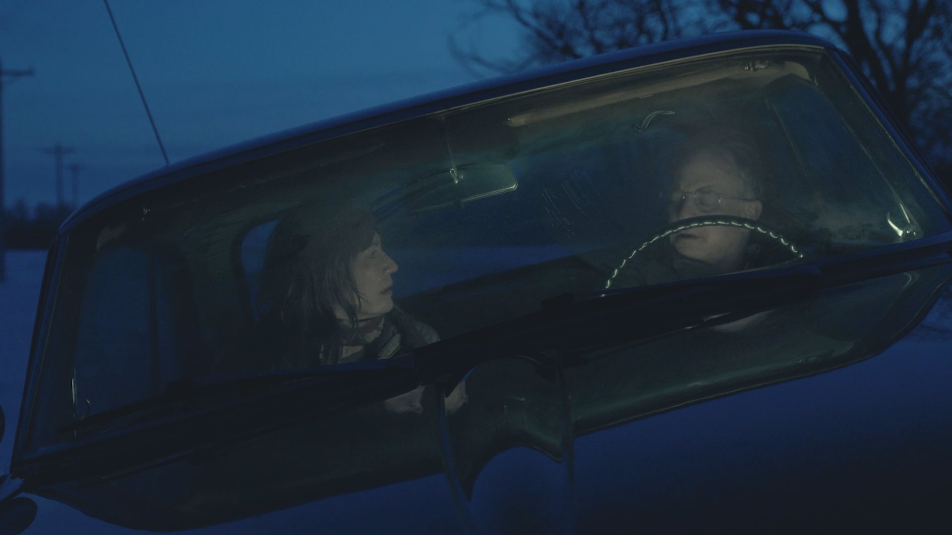 A man and woman sit in a car at night engaged in a serious and tense conversation. The scene highlights their expressions and interaction within the confined vehicle interior.