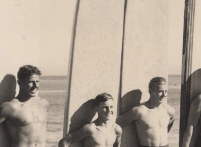 Three surfers pose beside their surfboards on a beach during a clear day. The scene transitions from a wide-angle view of the coastline to a close-up portrait of the individuals.