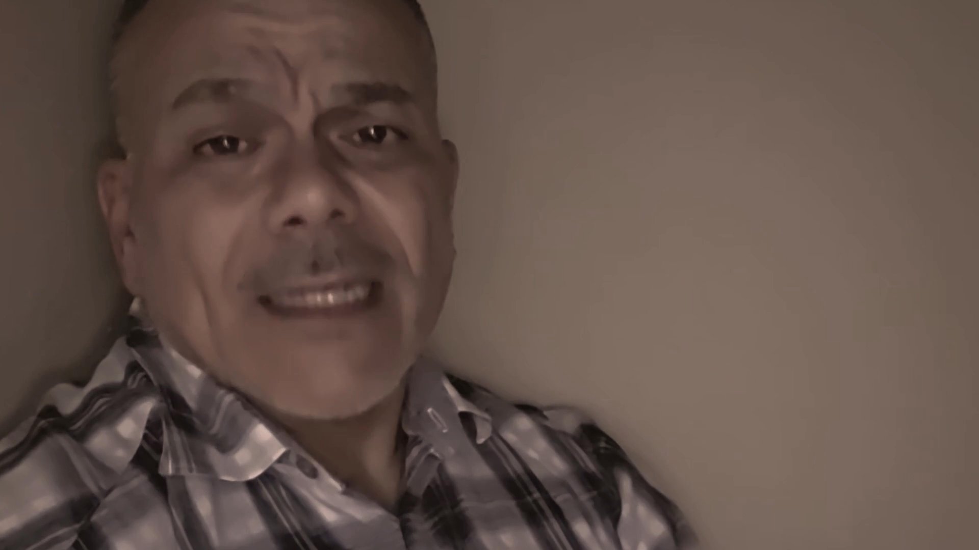 A man speaks directly to the camera, using varied facial expressions to convey his message during an indoor recording.