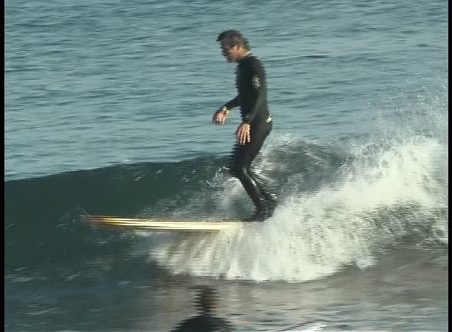 A surfer in a black wetsuit skillfully navigates an ocean wave on a longboard. The sequence captures the fluidity and balance maintained during the ride.