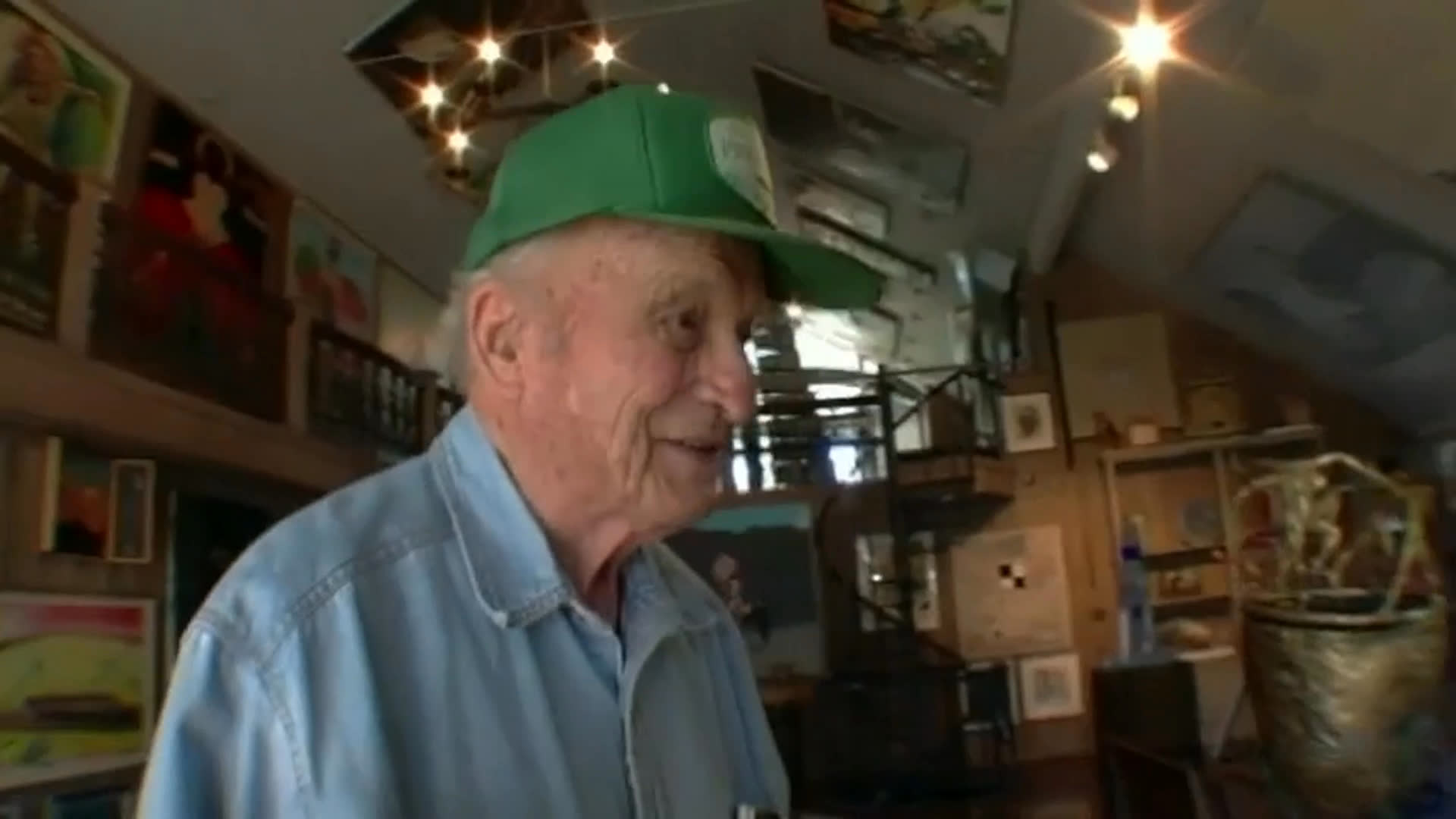 An elderly man in a green cap speaks among paintings, furniture, and collected objects in a densely decorated interior. The sequence cuts to a box truck beside vineyard rows, with captions noting that 50 works of art are departing on a two-year national tour.
