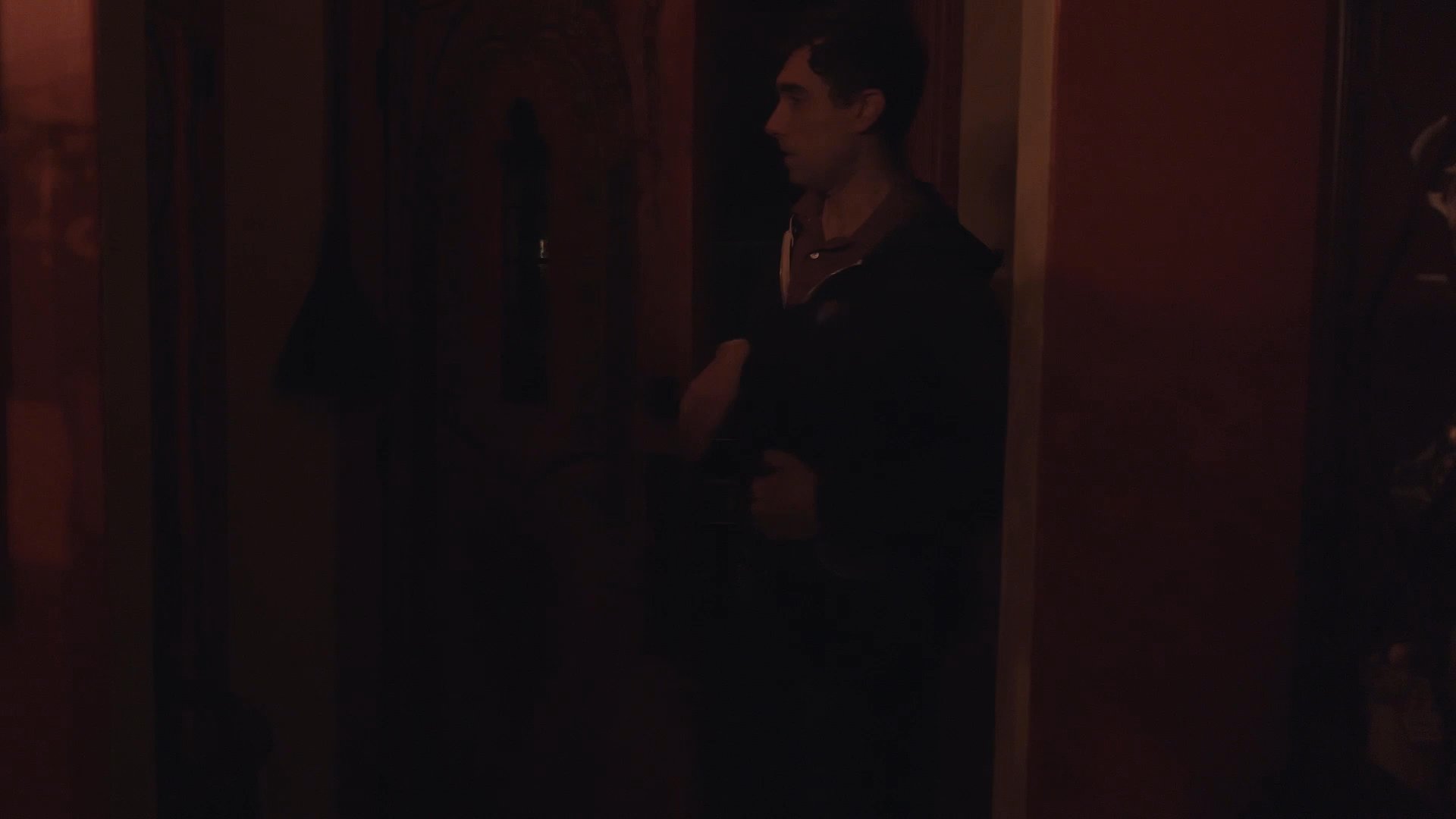 A man returns to a dimly lit home and encounters a woman sitting at a table. The scene captures a quiet, intimate moment between the two individuals in a domestic setting.