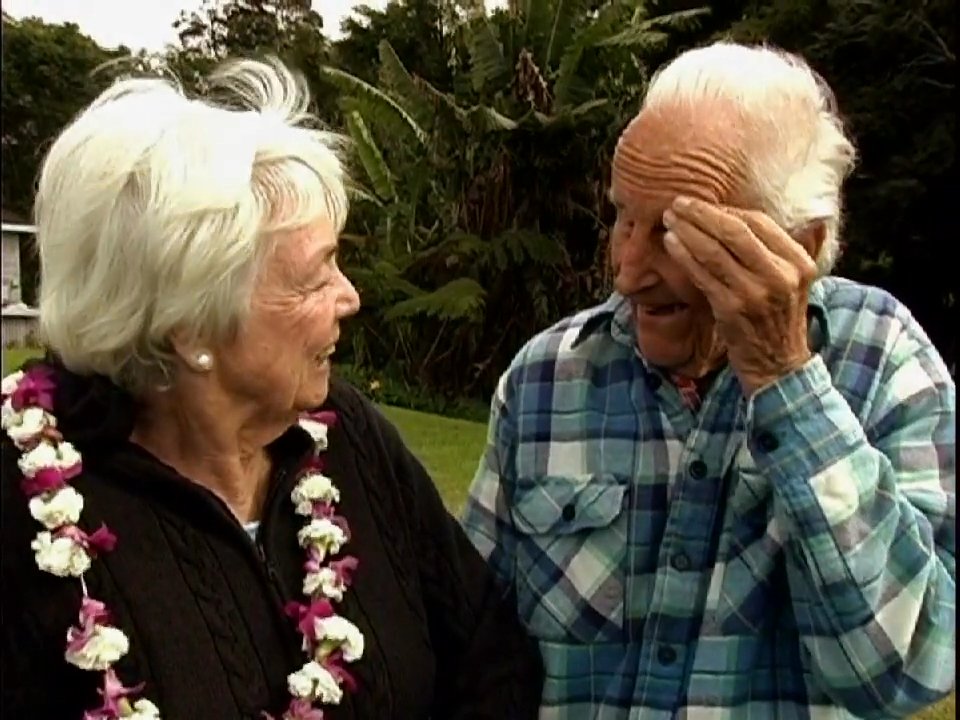 An elderly couple shares a conversation while wearing flower leis in a lush outdoor setting before joining a larger group for social interaction.