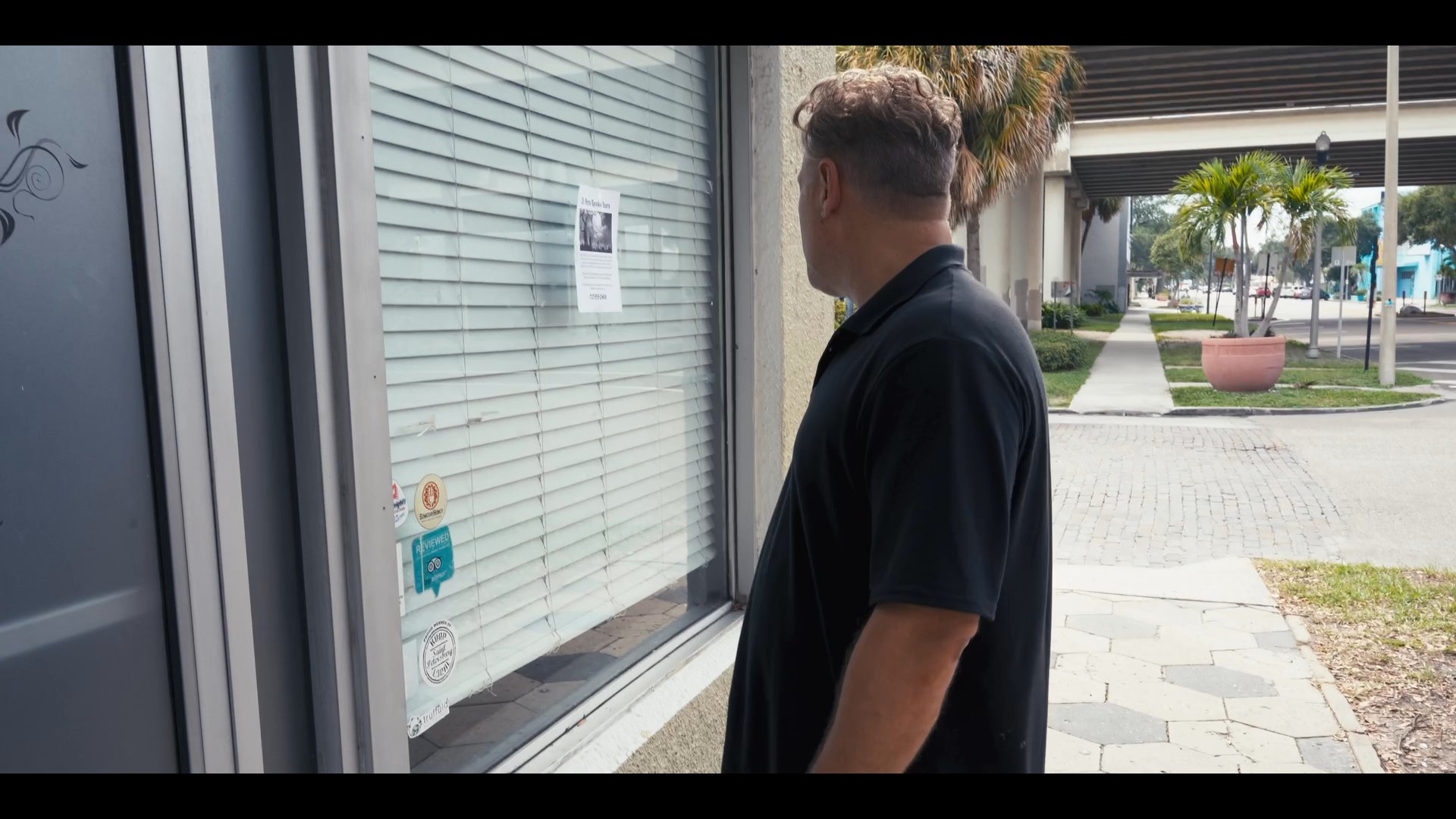 A man walks down a sidewalk and pauses to inspect a St. Pete Spooky Tours flyer posted on a shop window. He examines the advertisement closely while standing on the exterior of the urban storefront.