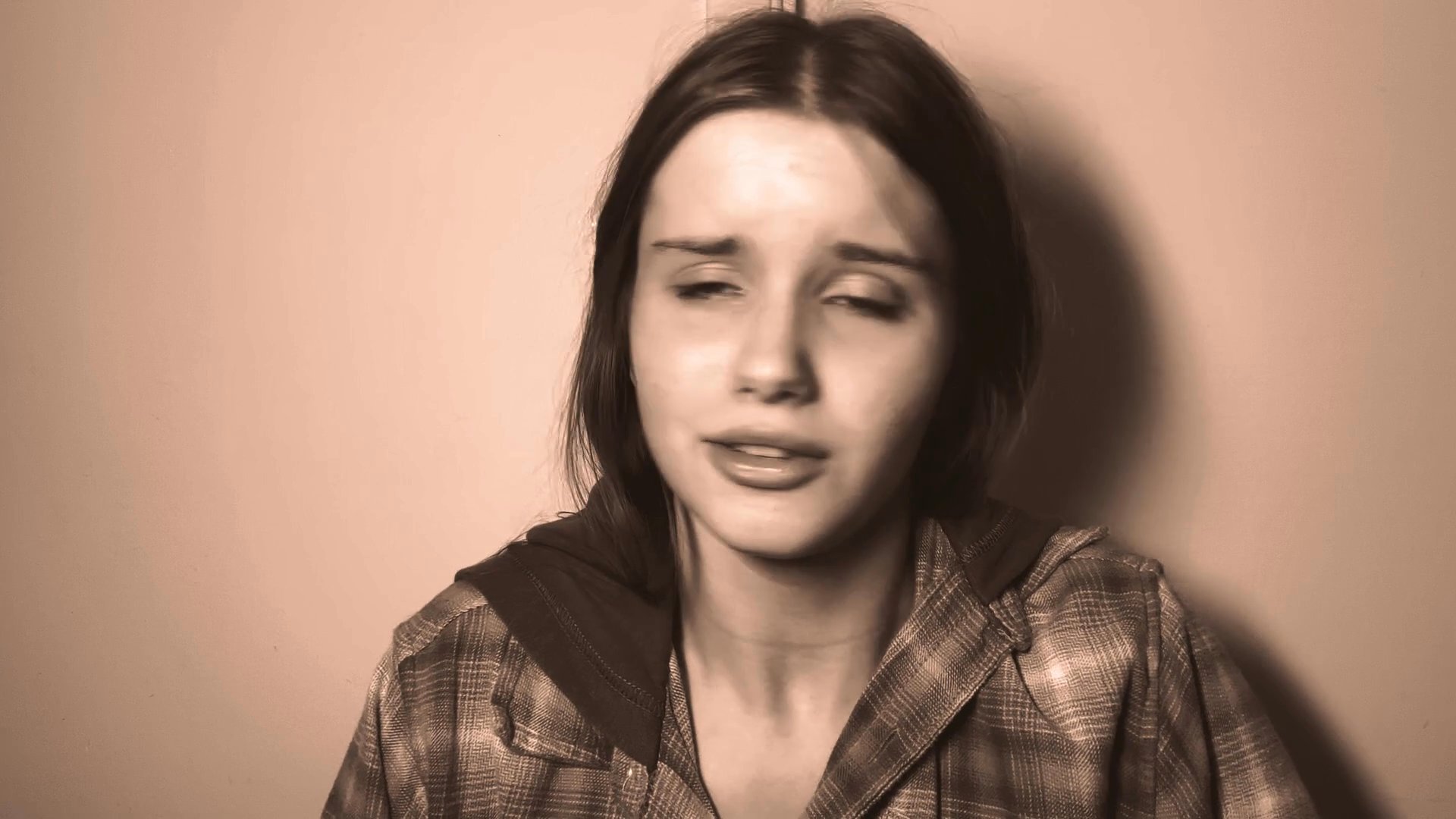 A young woman in a plaid shirt displays a range of intense, sorrowful expressions while speaking or singing against a plain background. The sepia-toned sequence captures a raw, intimate moment of emotional vulnerability.
