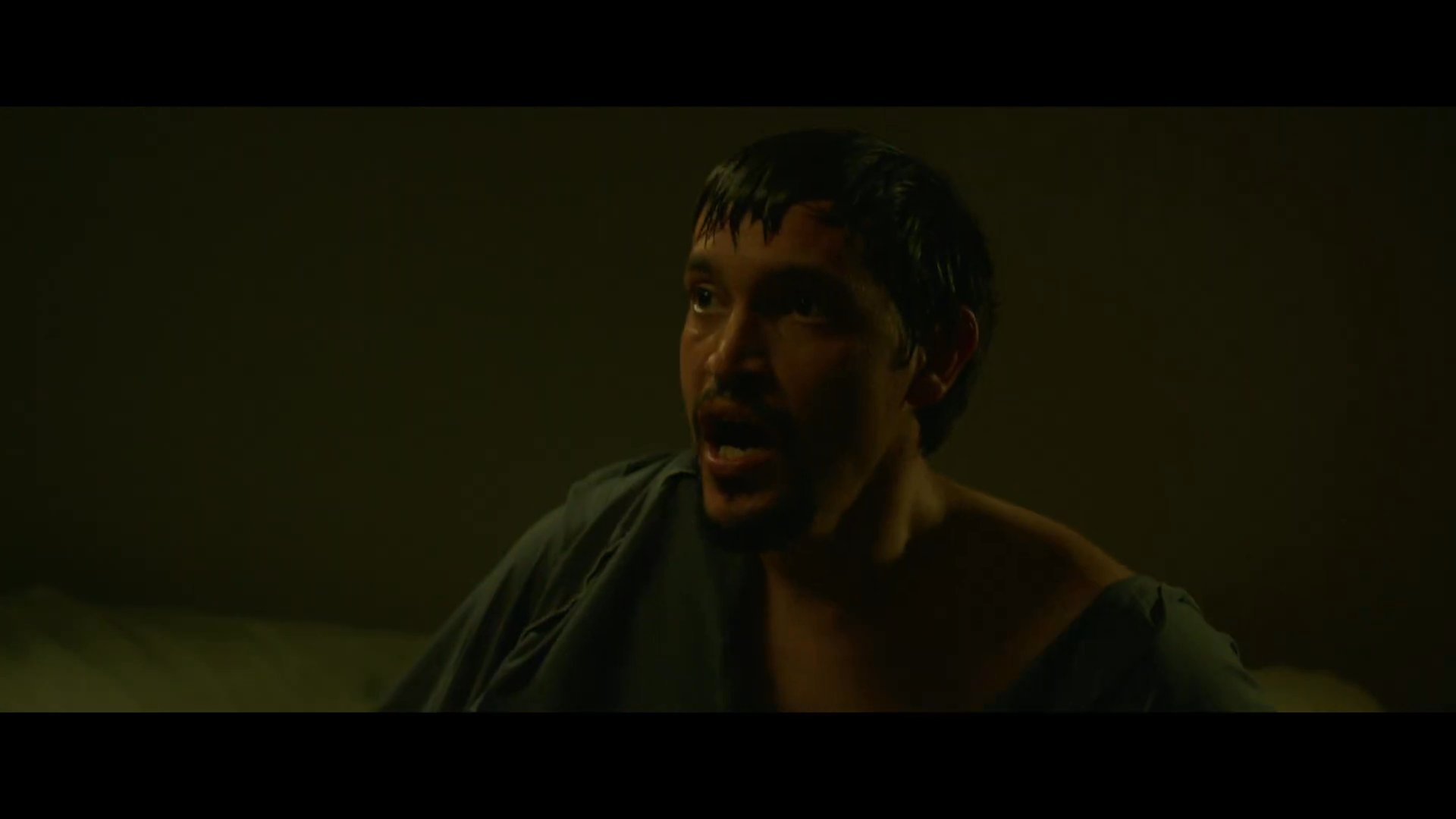 A man jolts awake in a dimly lit bedroom, displaying signs of distress and physical trauma as he confronts a disturbing reality.