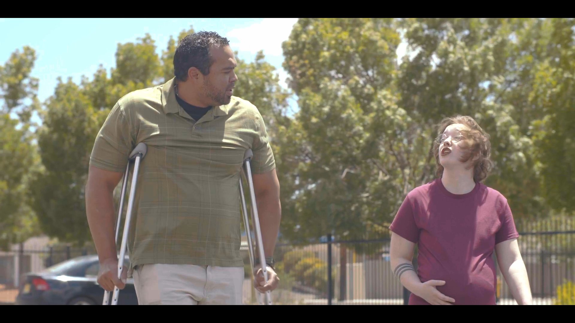 A man using crutches walks through an outdoor parking lot accompanied by a woman before they enter a vehicle. The scene highlights daily mobility support and transition into transportation.