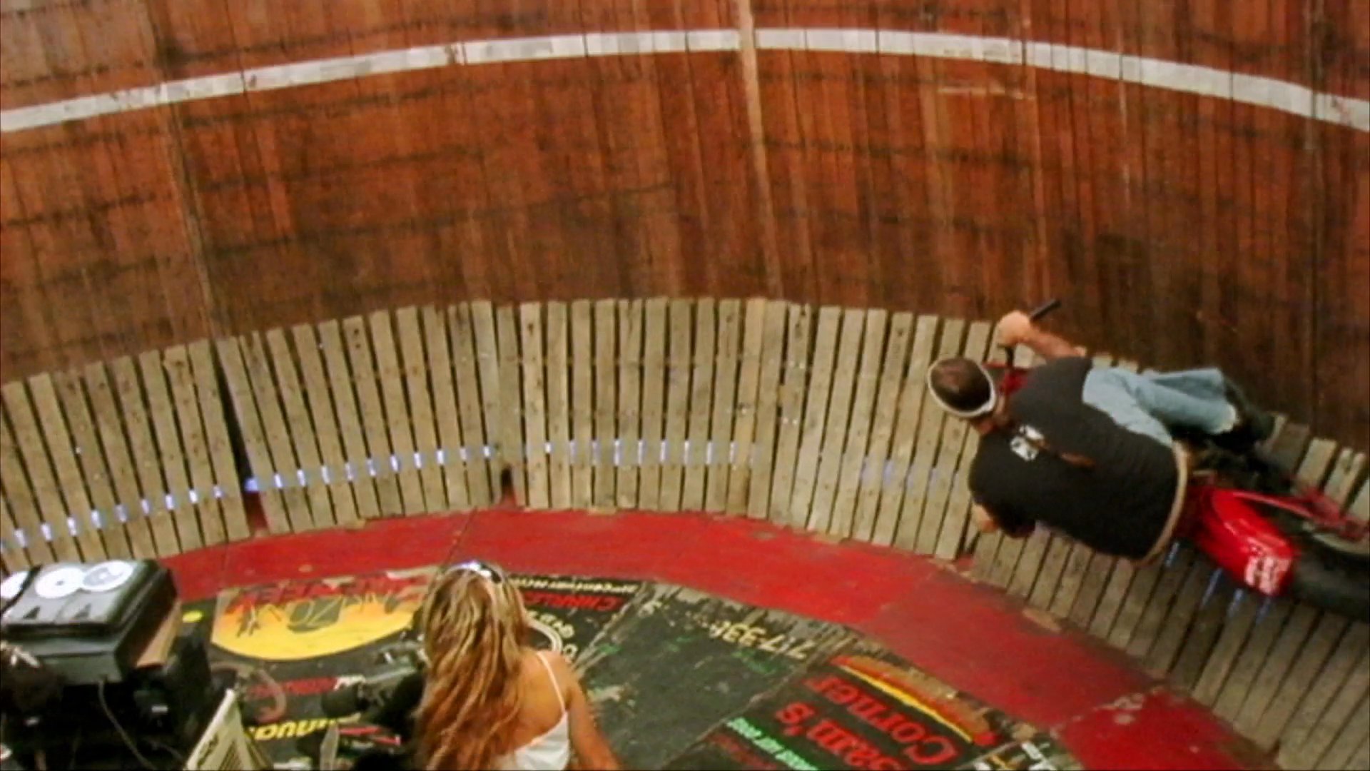 A motorcyclist performs high-speed stunts by riding along the vertical interior walls of a wooden cylinder. Spectators watch as the rider maneuvers the bike horizontally against the banked surface of the wall.