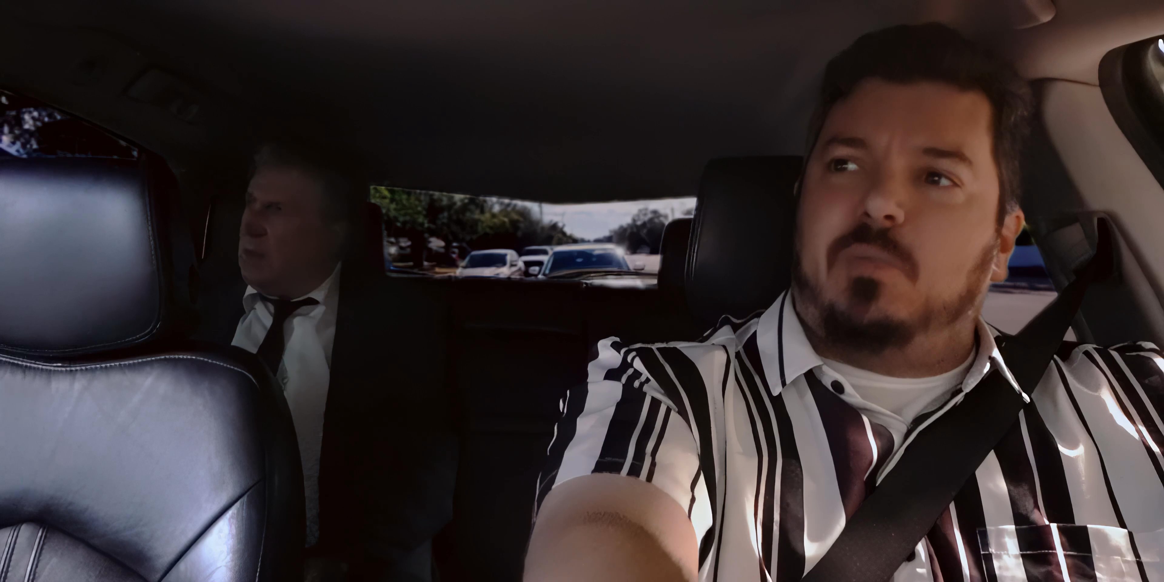 A man drives a vehicle while engaging in a serious conversation with a passenger seated behind him. The exchange involves direct eye contact through the rear-view mirror as they navigate the road.