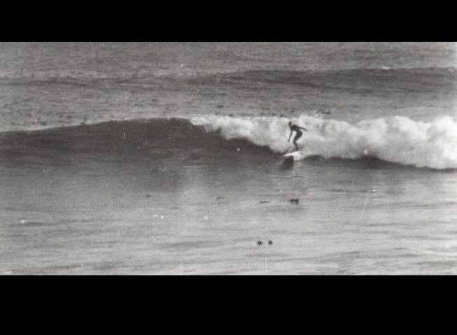 Vintage archival footage captures surfers at Cowell's Beach, featuring a posed group with a vehicle and a lifeguard standing by a tower. The sequence transitions between coastal portraits and individuals riding waves.
