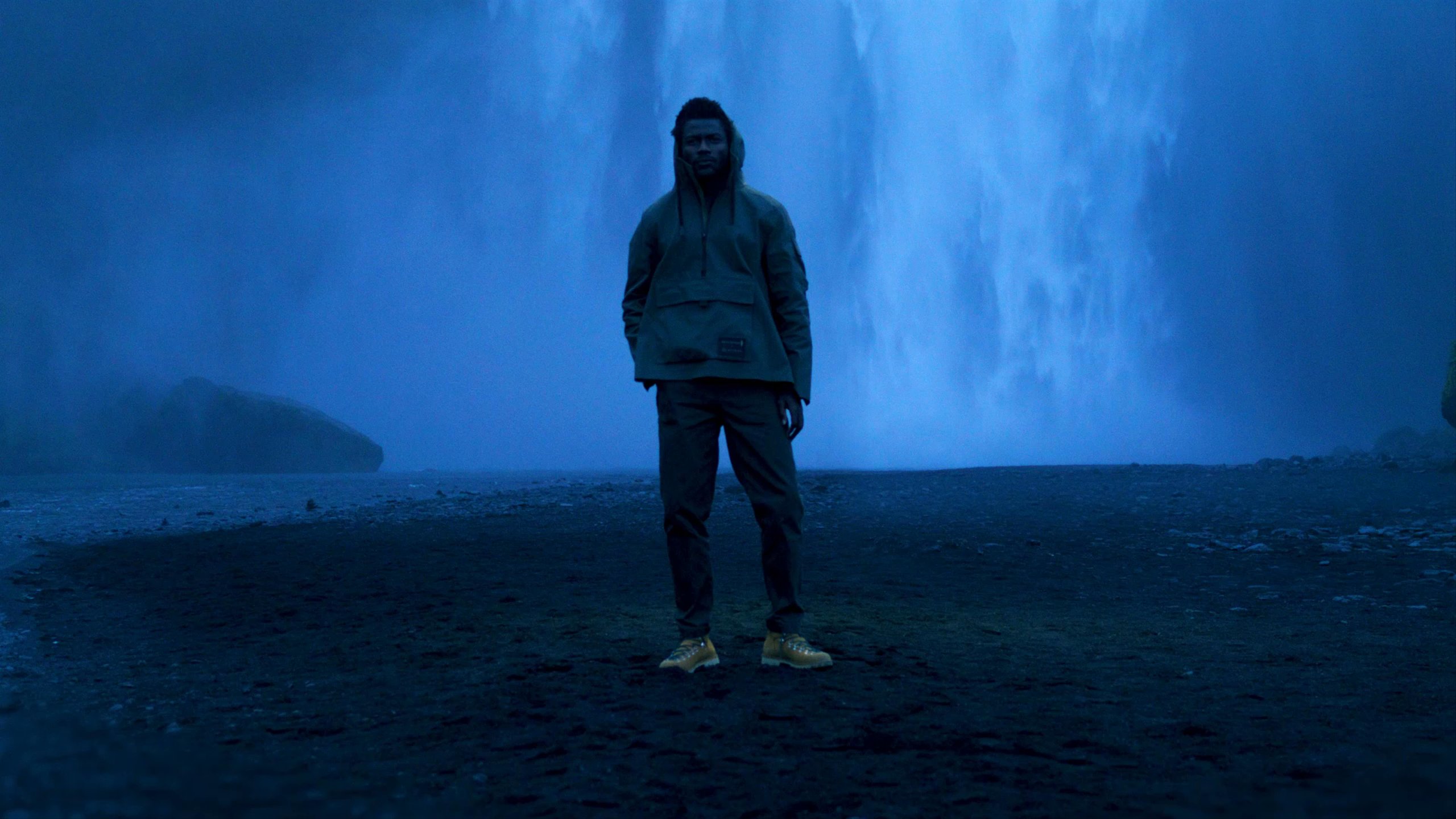Man wearing AETHER + Mackintosh Field Anorak standing in front of waterfall in Iceland