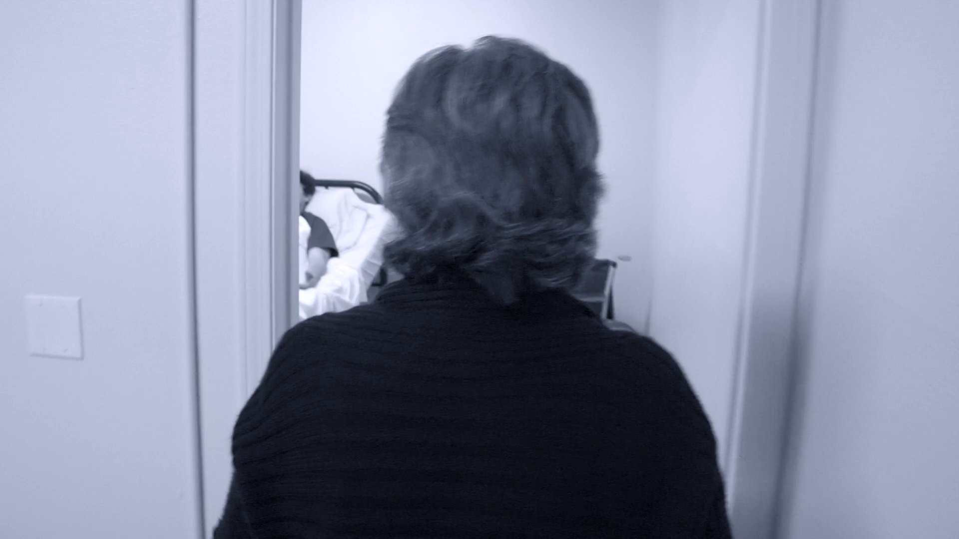 An older woman provides care and support to a man confined to a hospital bed while navigating the stresses of the situation. Her actions reflect a mixture of professional diligence and emotional concern in a medical environment.