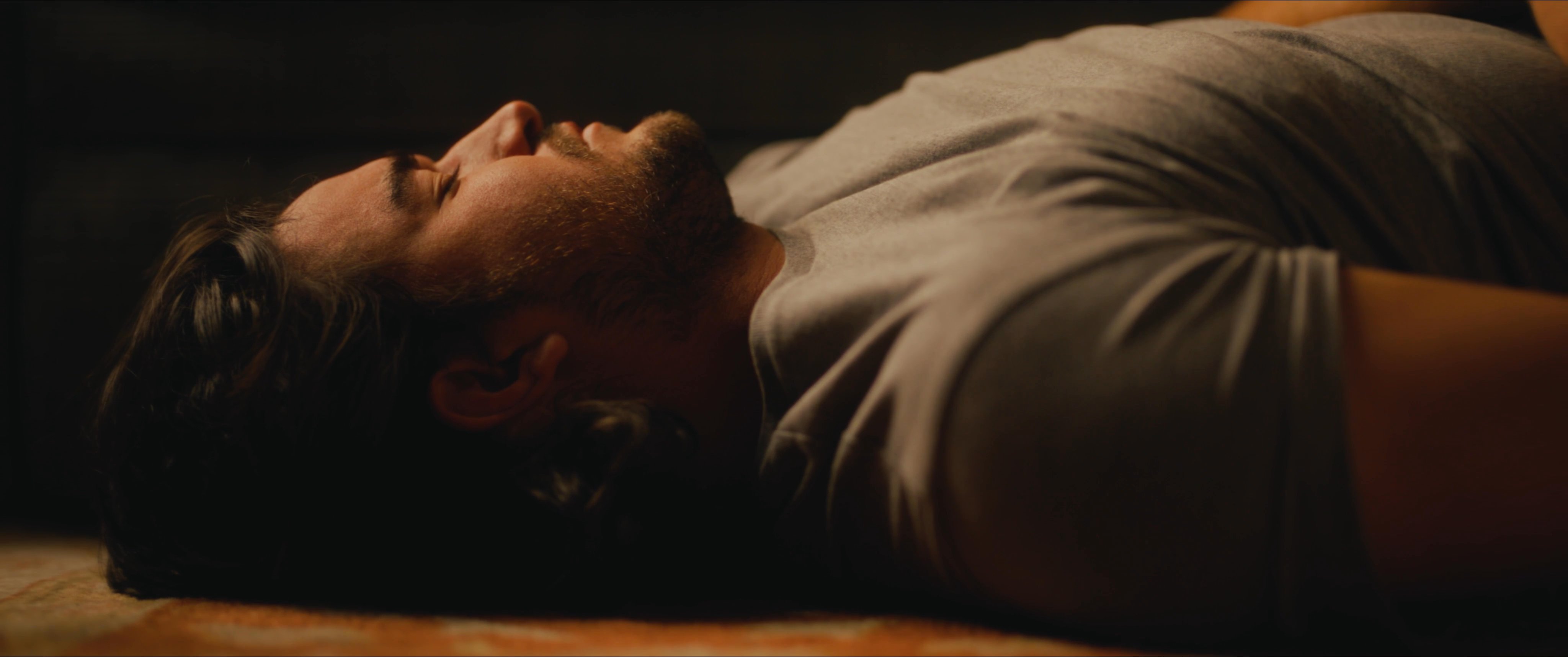 A man lies on the floor of a dimly lit, rustic cabin near several empty beer bottles. The atmosphere conveys a sense of stillness and isolation in the evening hours.