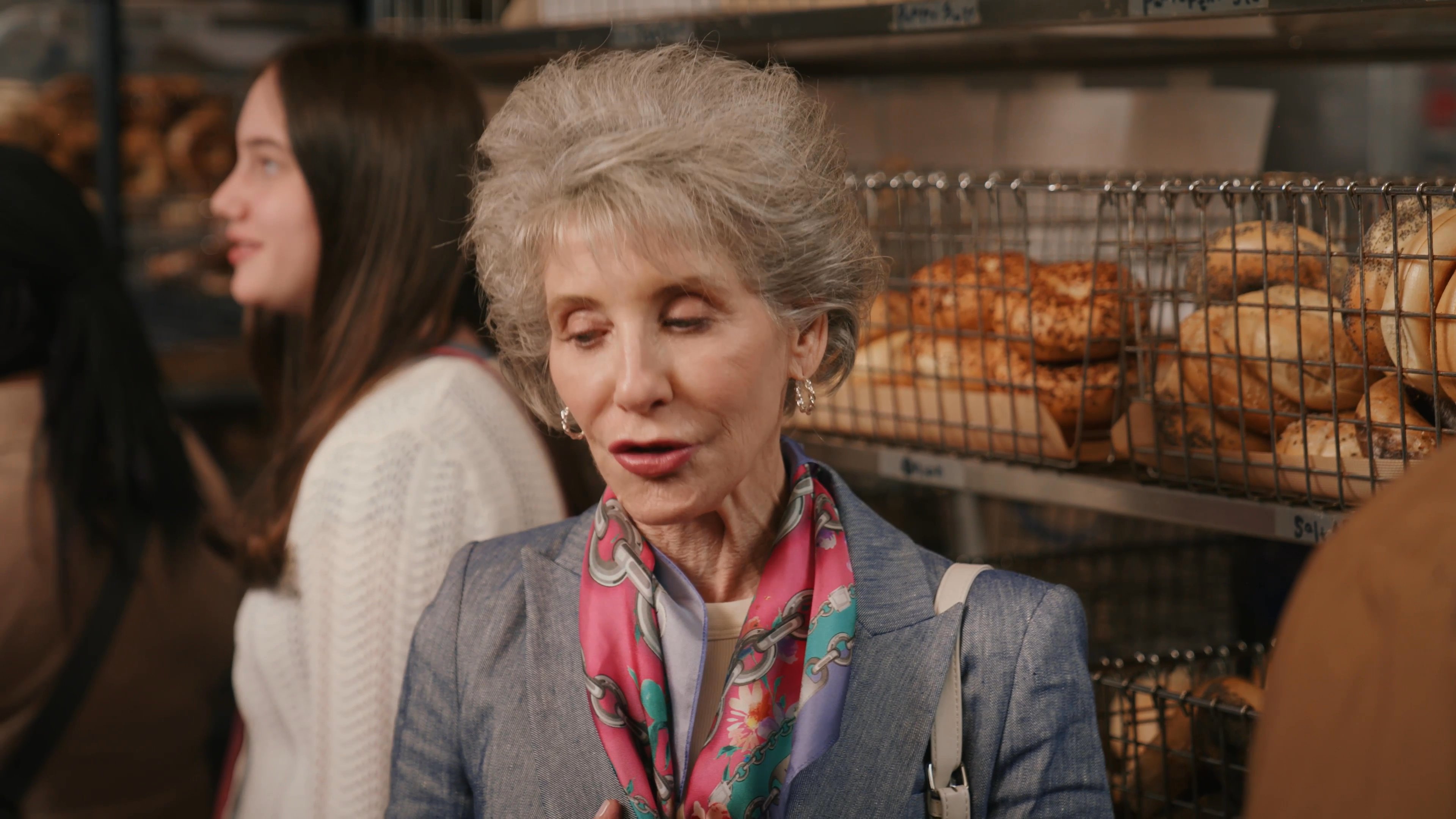 An elderly woman engages in an animated conversation with a man at a bakery counter. The two maintain a friendly dialogue while browsing the selection of baked goods.