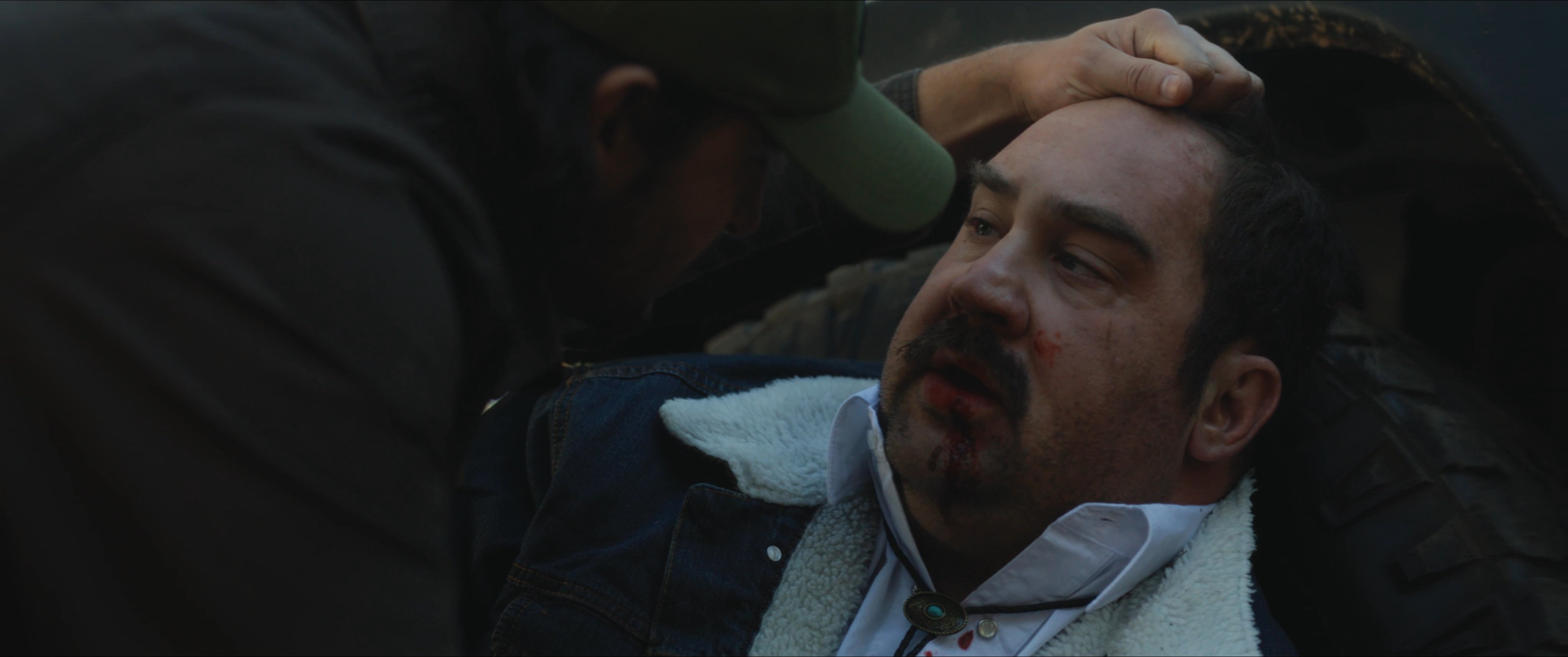 An injured sheriff sits against his patrol vehicle while an individual approaches him in a remote wooded area. The scene concludes with a wide shot of the sunlit forest floor where tactical equipment lies abandoned.