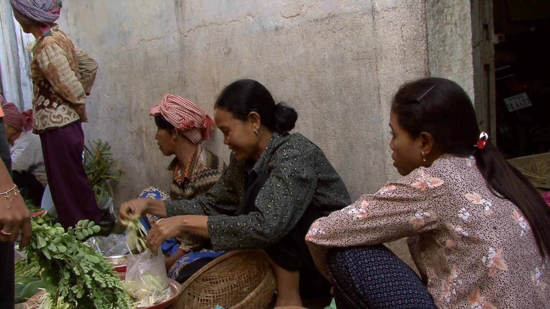 Locals engage in daily commerce at a bustling outdoor market, exchanging fresh produce and prepared goods. Vendors and customers interact within the vibrant, stall-lined atmosphere.