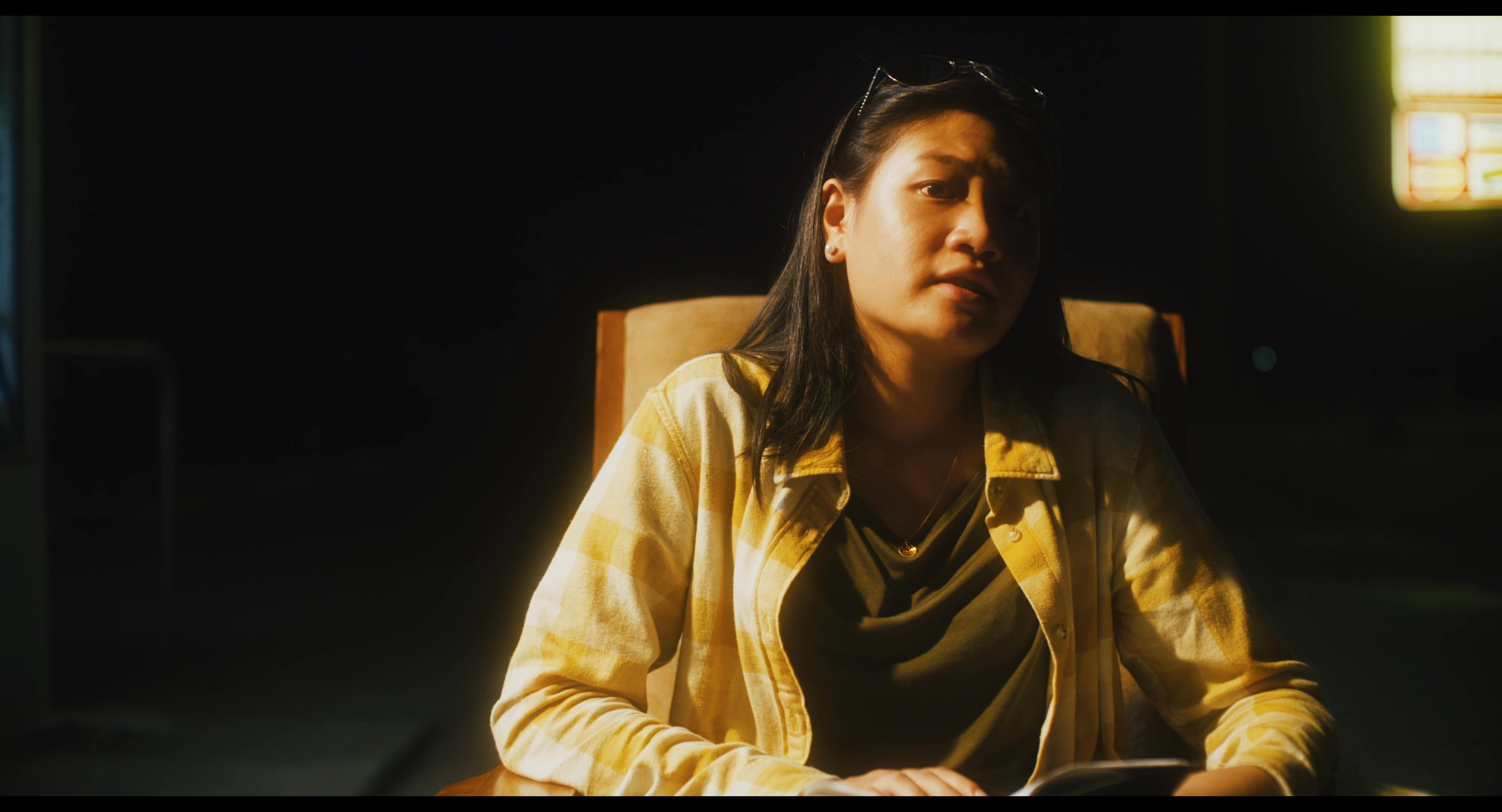 A woman sits in a warmly lit, shadowed room while engaging in a conversation. The scene transitions to include a man listening, highlighting the exchange between two individuals.