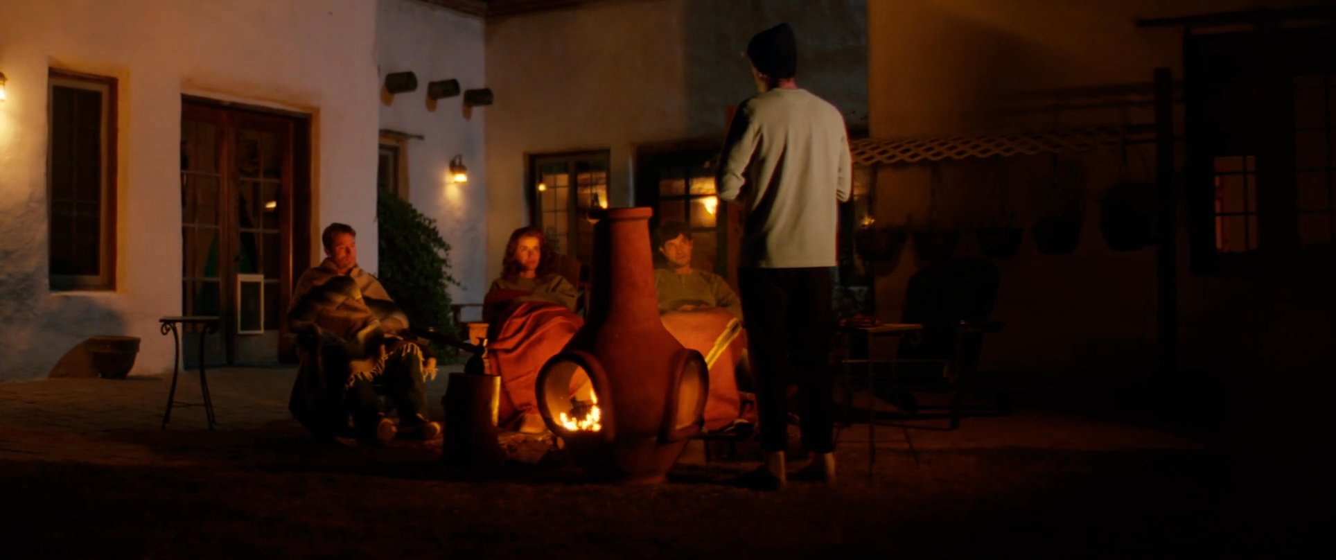 A group of people gathers around an outdoor fireplace in a dimly lit courtyard for an evening conversation. The warm glow of the fire illuminates the subjects as they engage in a shared discussion.
