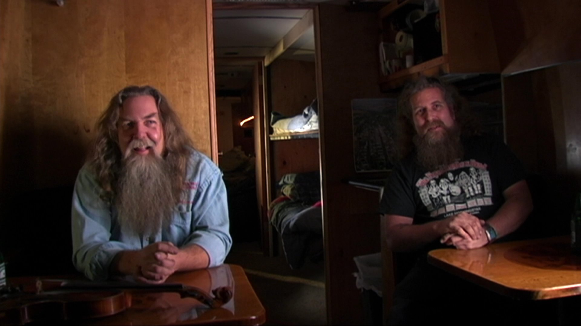 Two bearded musicians perform an acoustic jam session, transitioning from technical instrument adjustments to a seated dialogue and collaborative musical exchange.