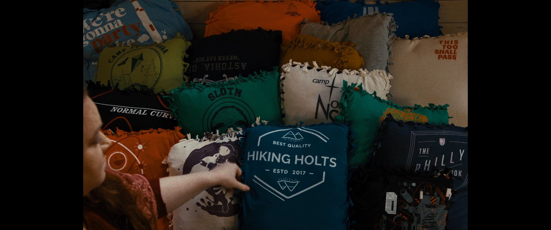 An individual organizes a collection of decorative cushions on a seating area within a rustic, wood-paneled room. The process transforms the space, culminating in a styled arrangement before the person captures a photo with a smartphone.