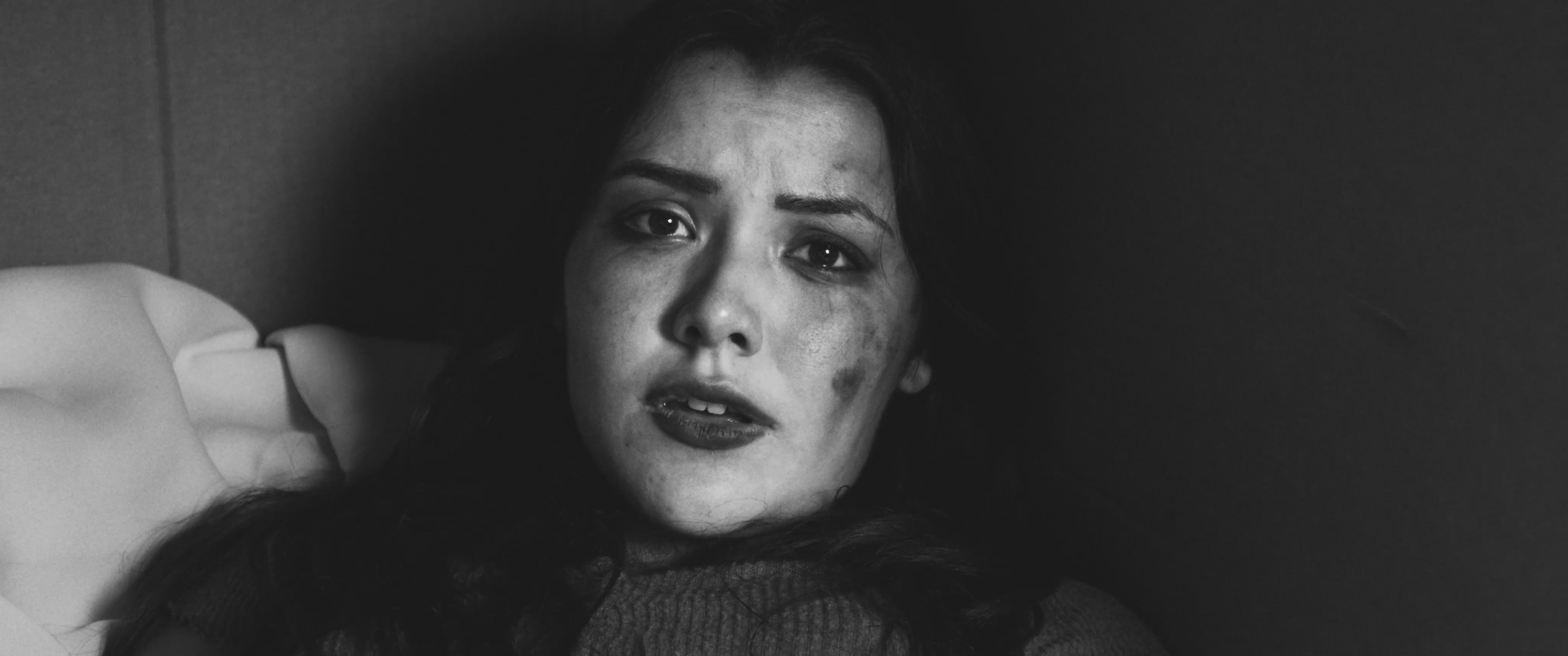 A woman with a distressed expression stares directly into the camera in a moody, low-light setting. The black-and-white visual highlights her somber mood and internal struggle.
