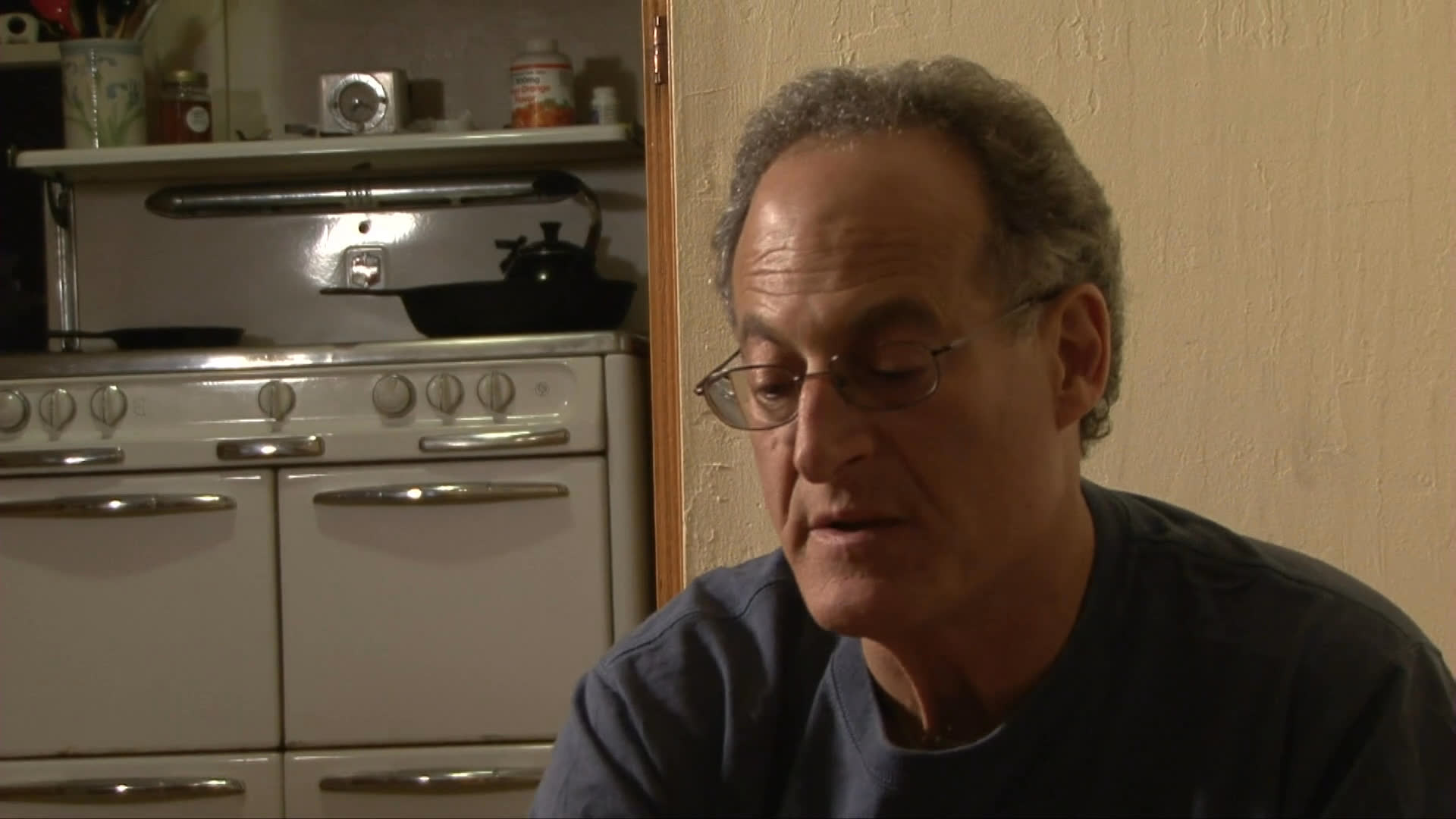A man speaks while inside a kitchen, alternating with scenes of a black dog transitioning from the outdoors to resting quietly on a rug indoors.